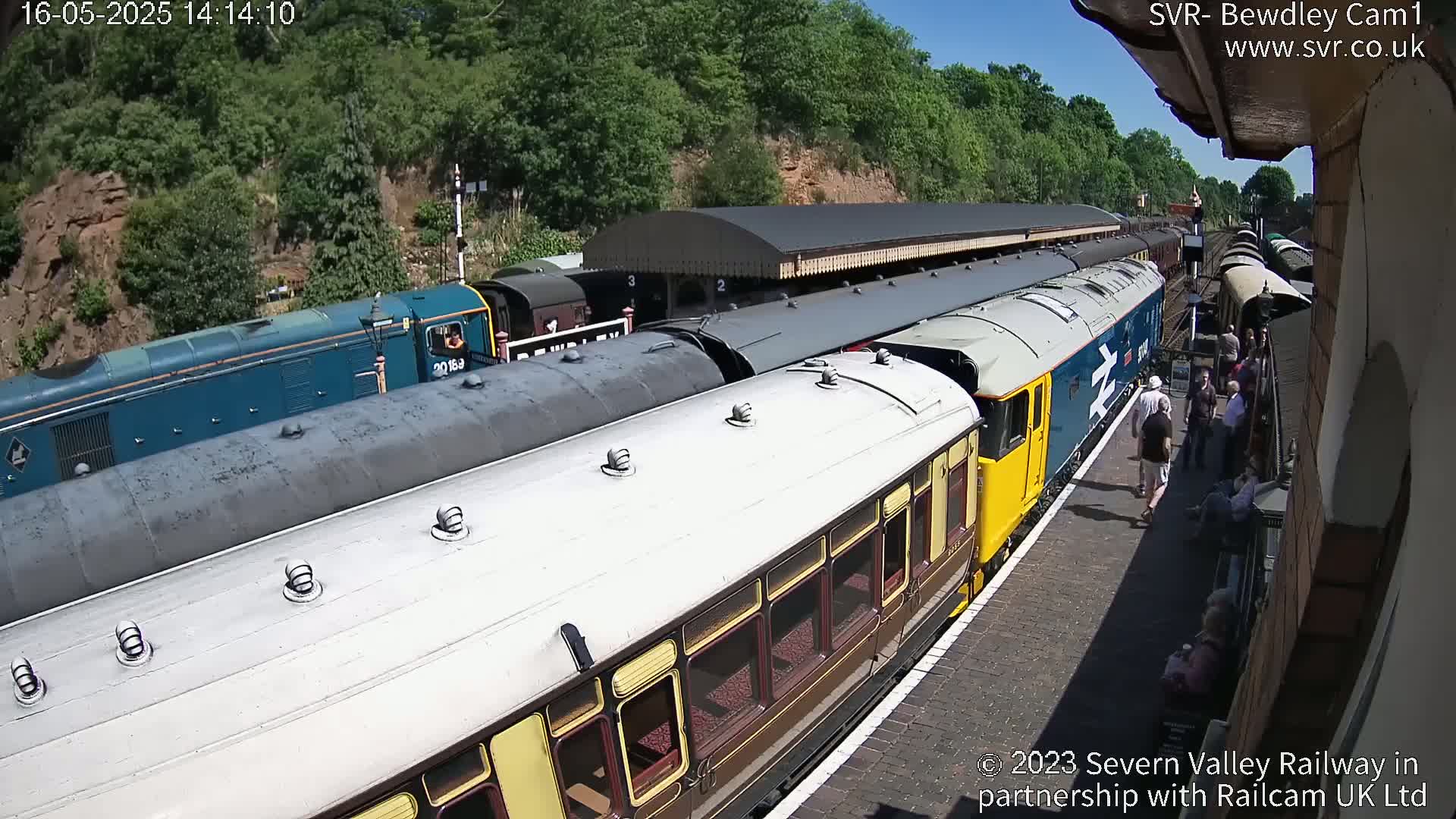 A passenger train with brown and cream cars sits at a station platform on a sunny day, with several people standing nearby and additional train cars and a blue diesel locomotive visible in the background.