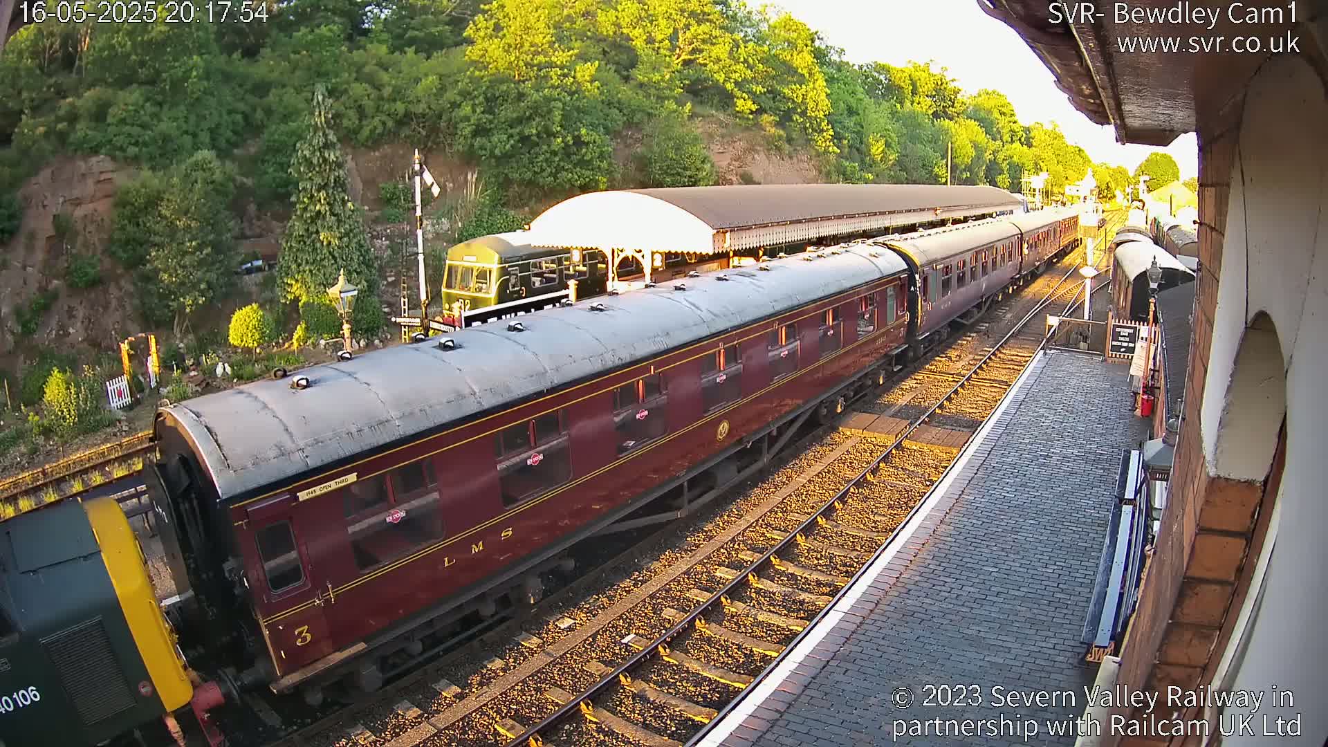 A maroon passenger train is departing a station at sunset, alongside a green diesel locomotive.