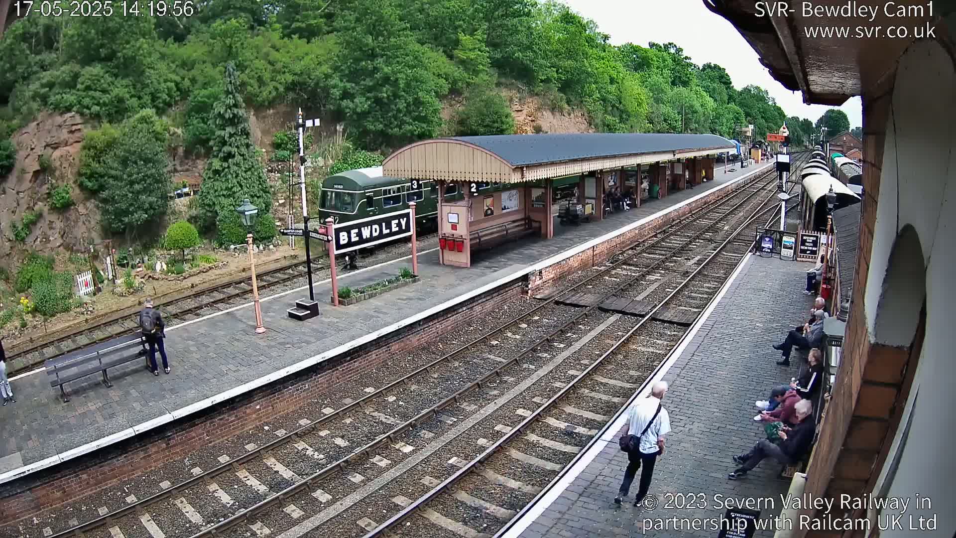 A train station platform with a dark-green train and several people, situated alongside train tracks that curve gently away from the viewer under a partly sunny sky.