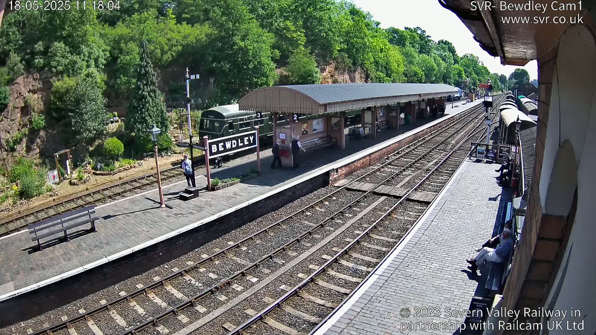 A sunny day at a train station with a green train parked at the platform, several people are present.