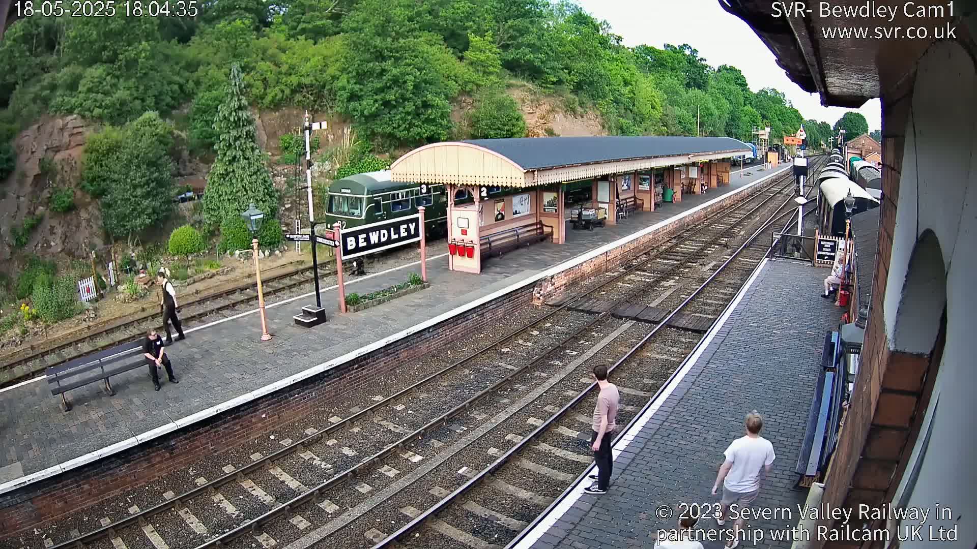 A train sits at a railway station platform on a partly cloudy day, with several people standing and sitting nearby.