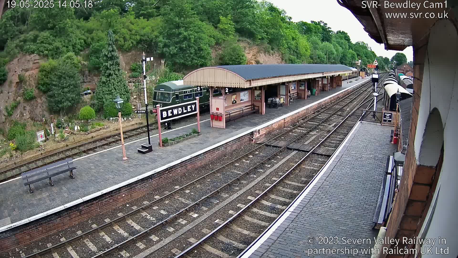 A train sits at a station platform on a partly sunny day, next to a covered waiting area and several railway tracks.