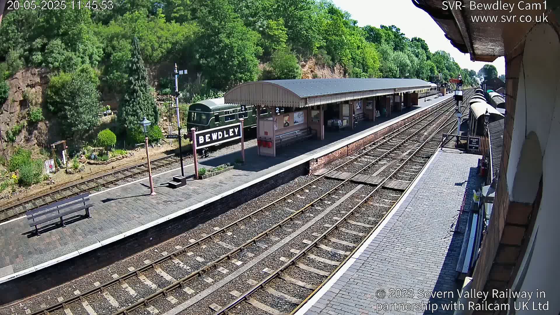 A train sits at a station platform on a sunny day, surrounded by lush green trees and a brick building.