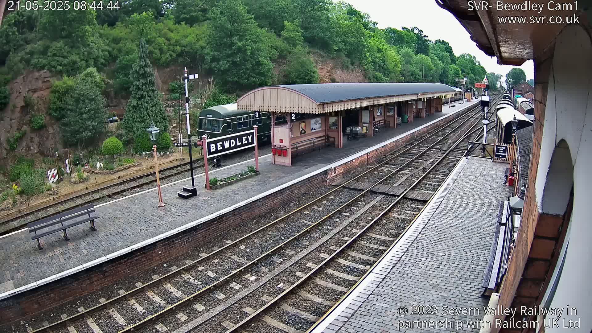 A train is stopped at a station platform next to a covered waiting area, under an overcast sky.