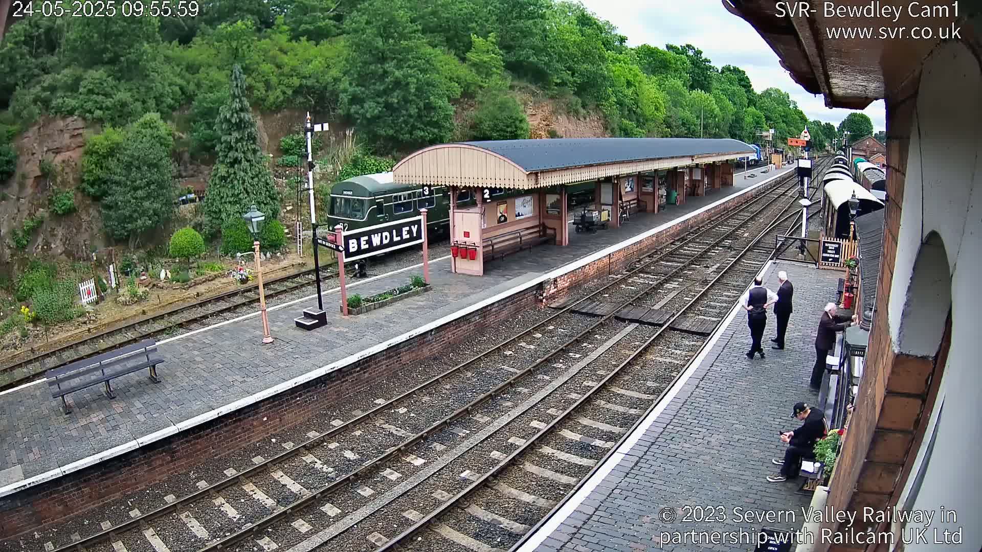 A train sits at a station platform next to a covered waiting area, with several people standing and sitting on the platform under partly cloudy skies.