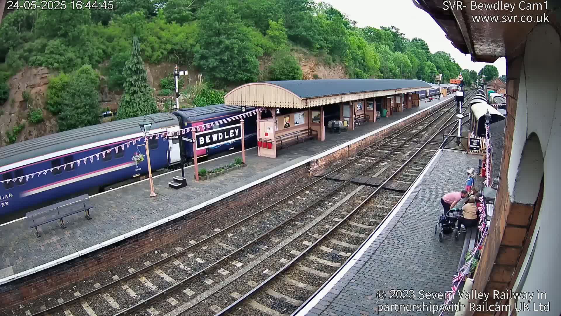 A passenger train is stopped at a station platform on a sunny day, with several people visible near a building and a wheelchair.