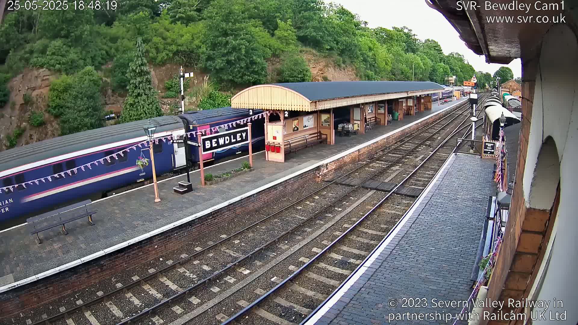 A train is stopped at a railway station platform next to a covered waiting area, with trees and a hillside visible in the background.