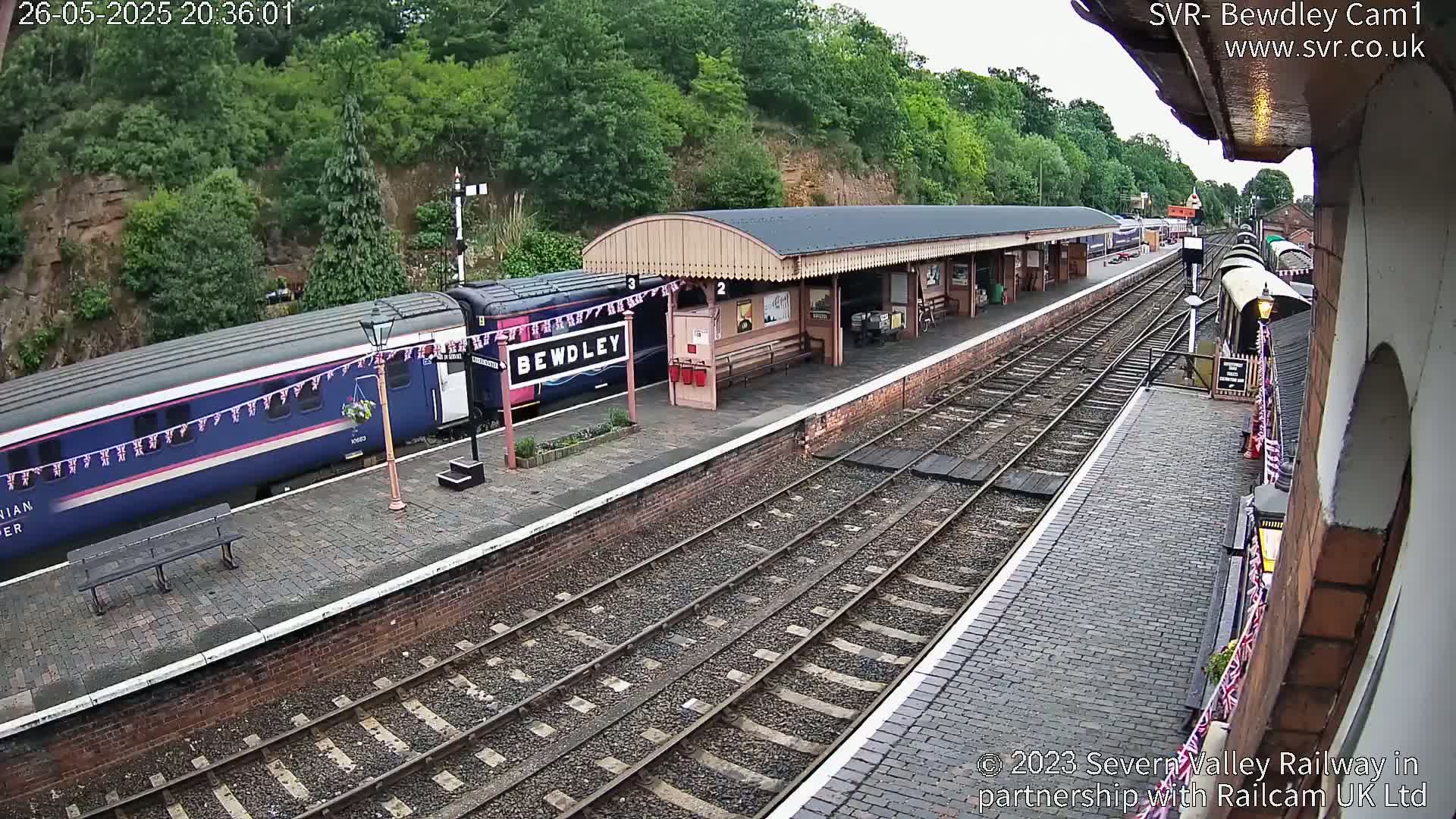 A blue passenger train is stopped at a railway station platform under a partly cloudy sky, with another train visible further down the tracks.