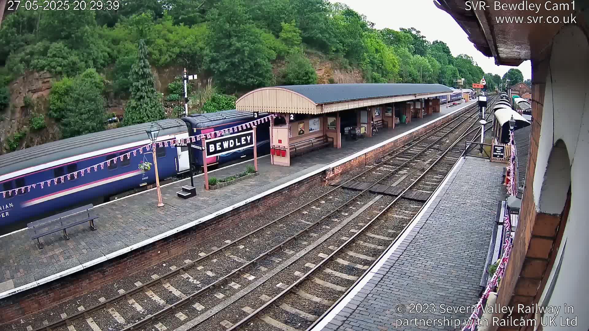 A train is at a station platform on a sunny day, with additional train cars visible further down the tracks.