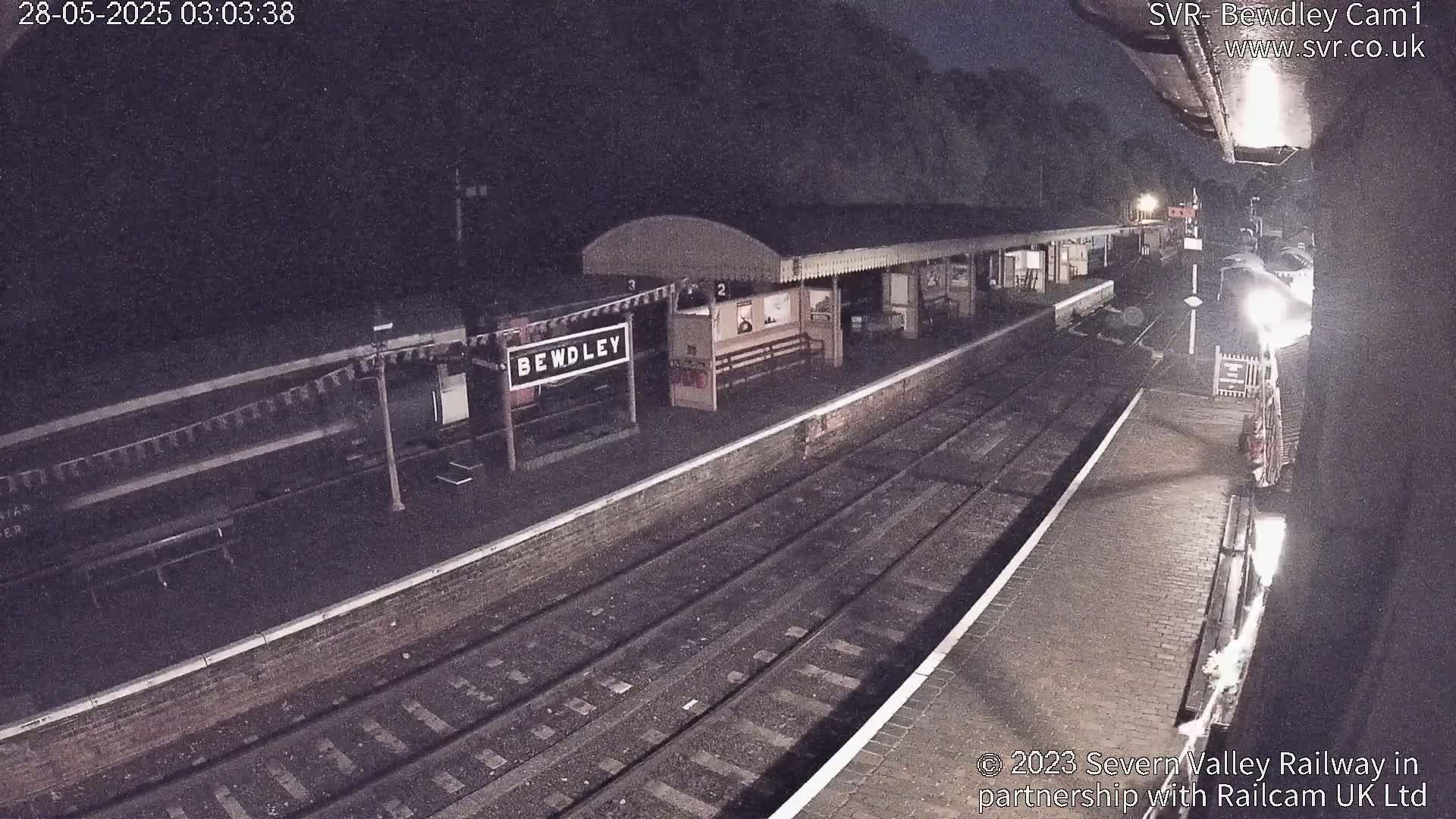 A nighttime view of a train station platform with train cars and tracks under dark, possibly overcast, conditions.