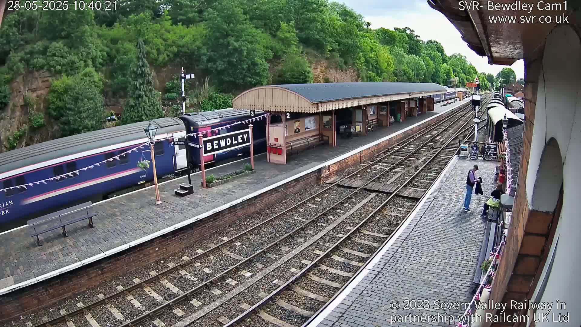 A blue and black passenger train is stopped at a railway station platform on a partly sunny day.