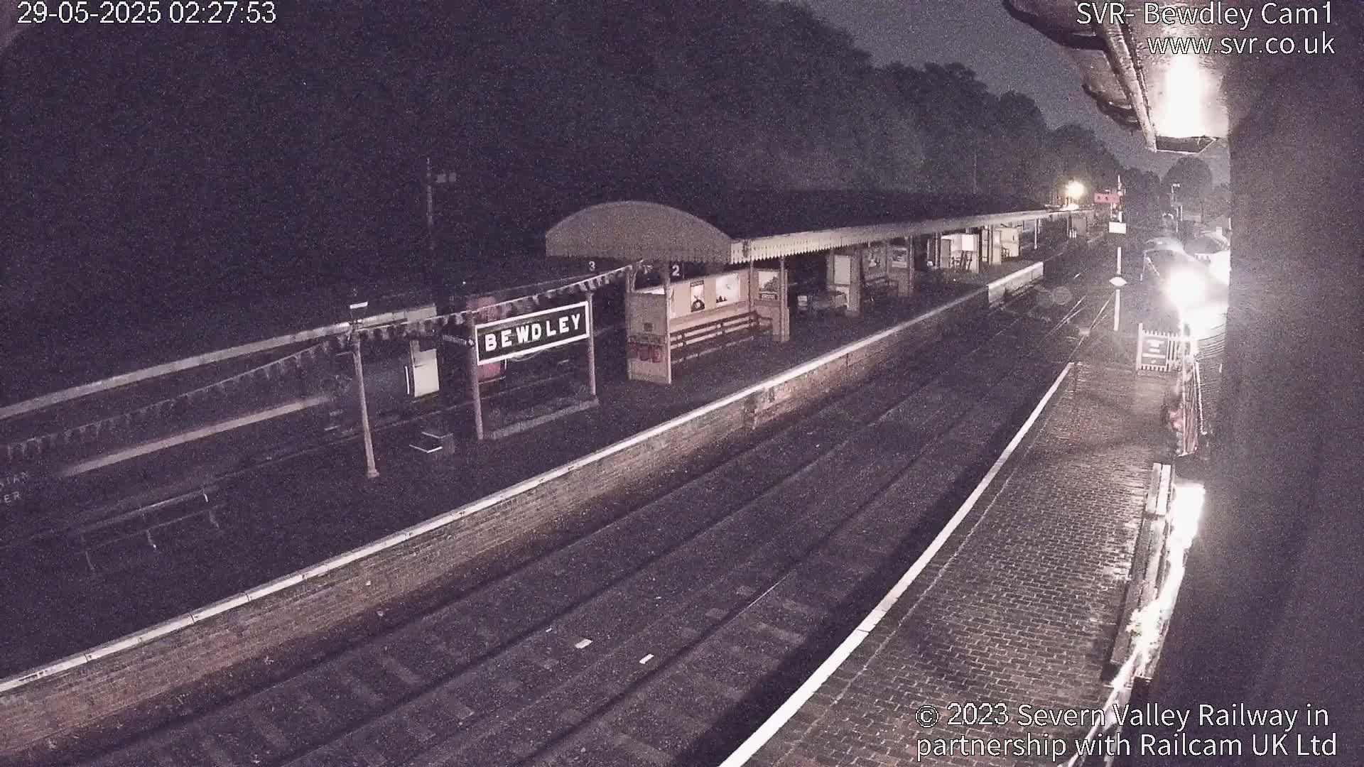 A nighttime view of a train station platform with a train partially visible, under dark, possibly rainy conditions.