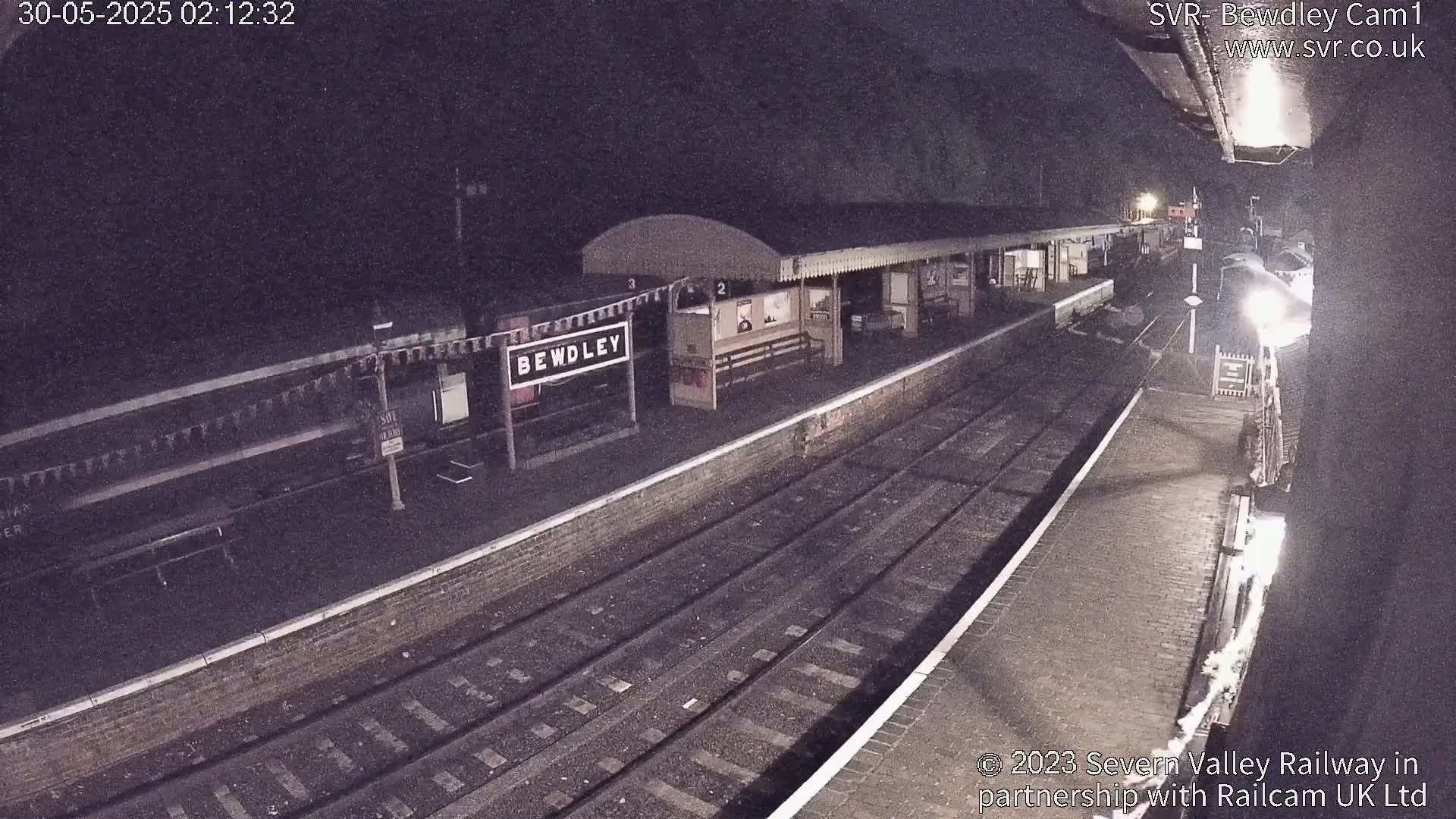 A nighttime view of a train station platform with train tracks and a partially visible train under dark skies.