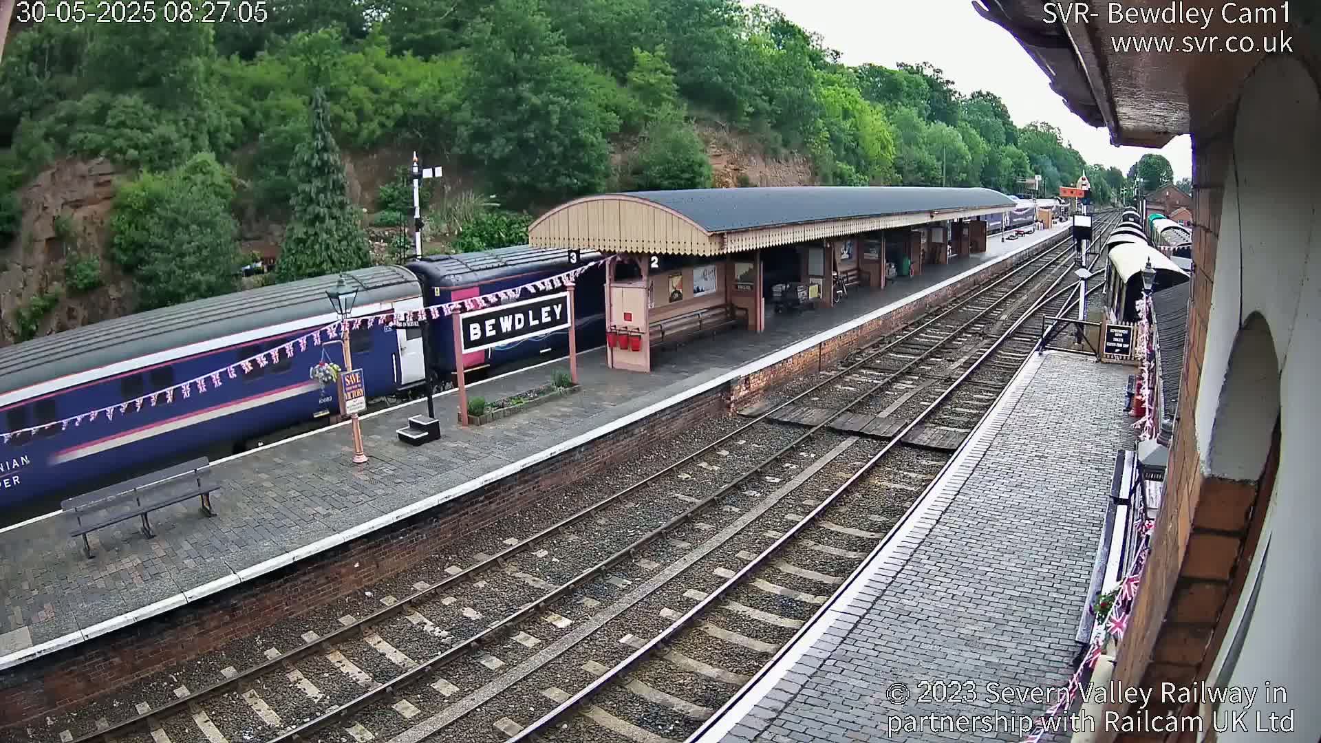 A train is stopped at a station platform on a partly cloudy day, next to a covered waiting area.
