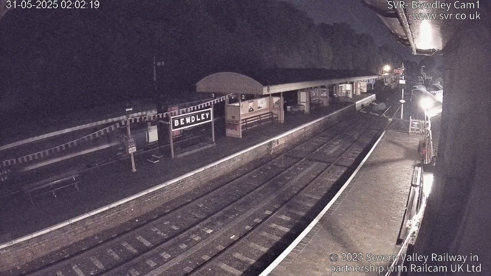 A nighttime view of a train station platform with train tracks and a partially visible train.