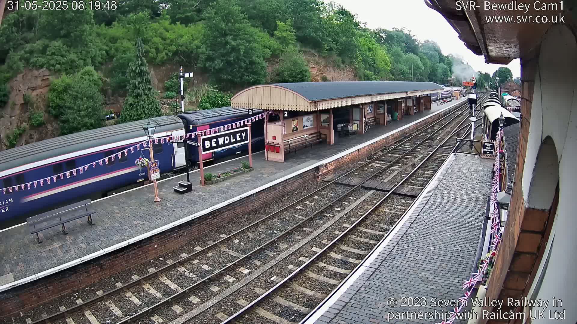 A train is at a station platform on a partly sunny day, with additional train cars visible further down the tracks.