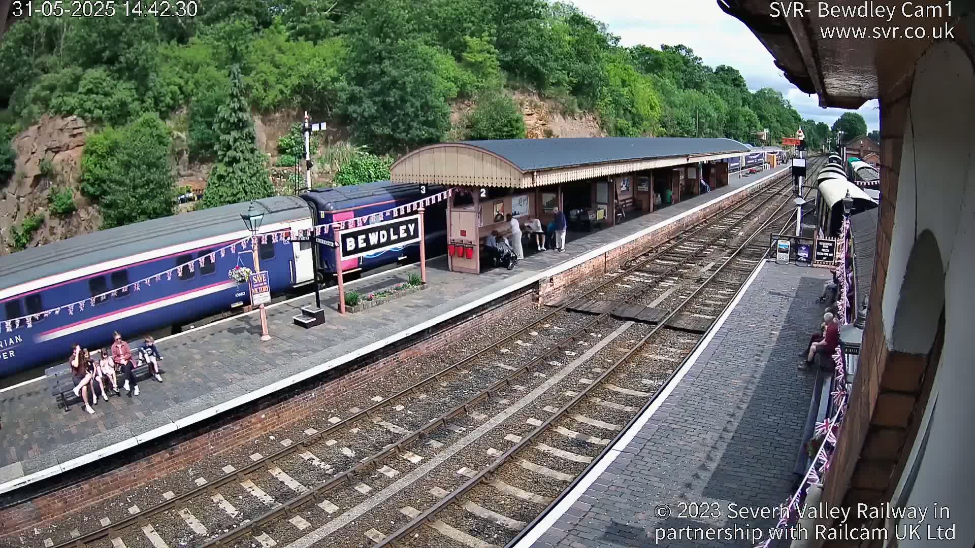 A train is stopped at a station platform on a sunny day, with several people waiting on the platform and others seated on a bench.
