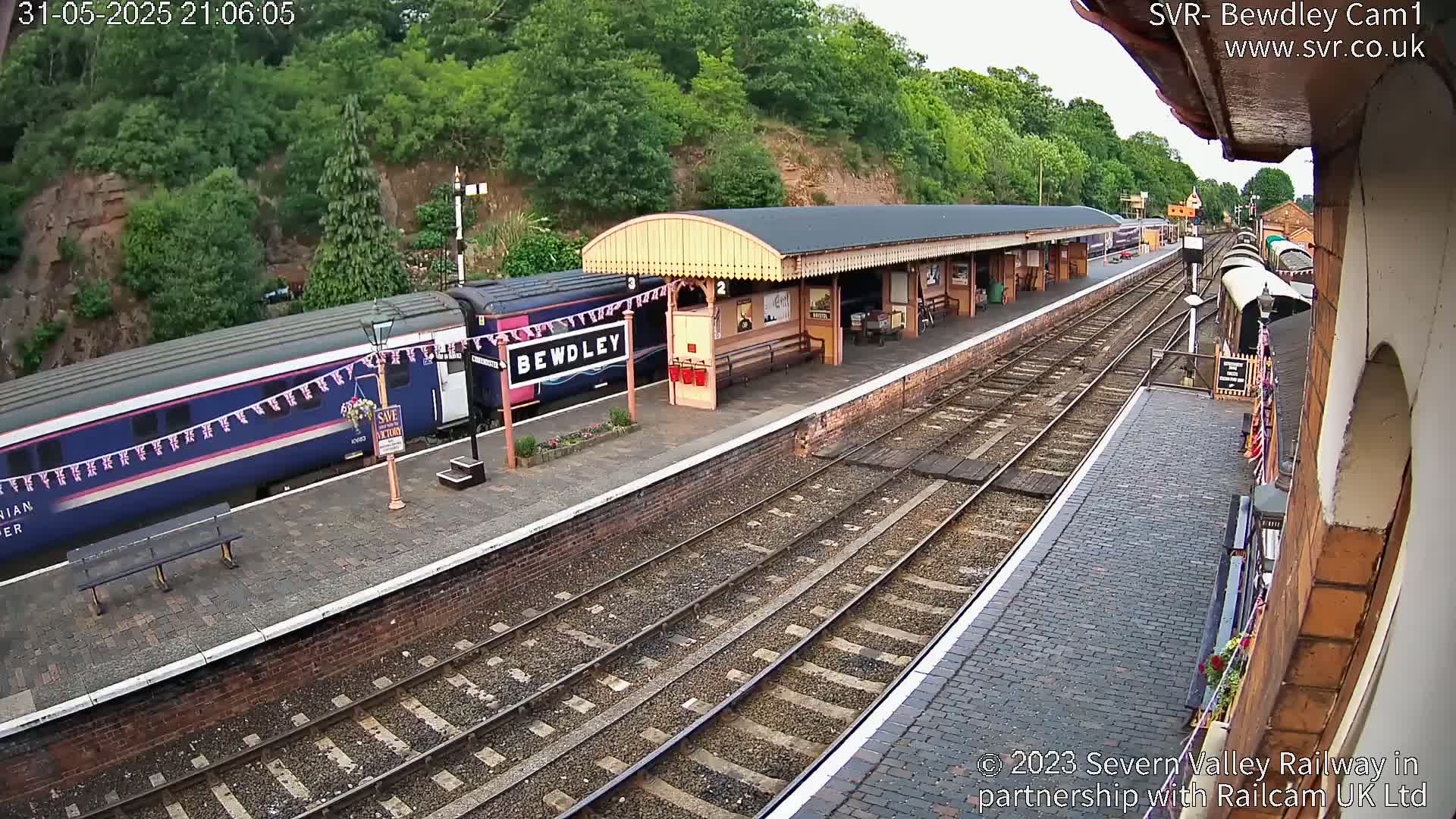 A train is stopped at a station platform next to a covered waiting area, with a backdrop of trees and hills under a clear sky.