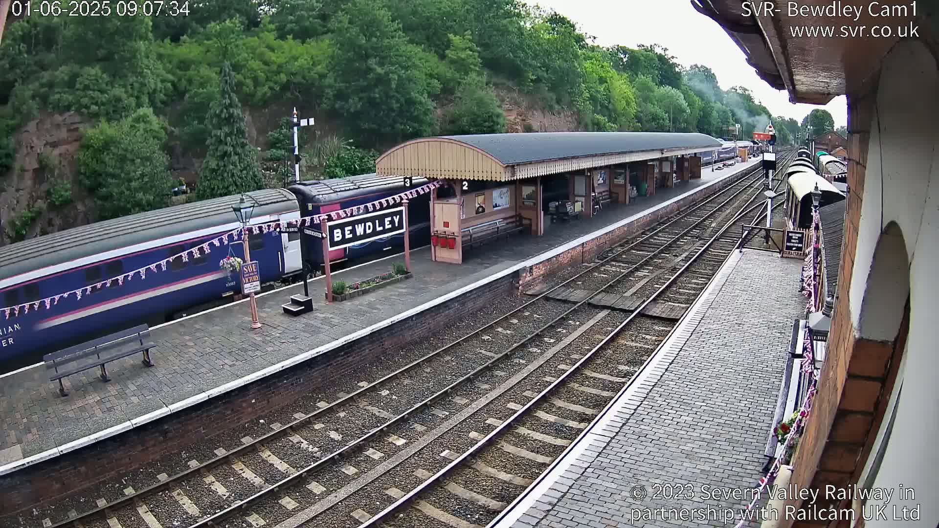 A train is at a station platform on a partly cloudy day, with additional trains visible further down the tracks.