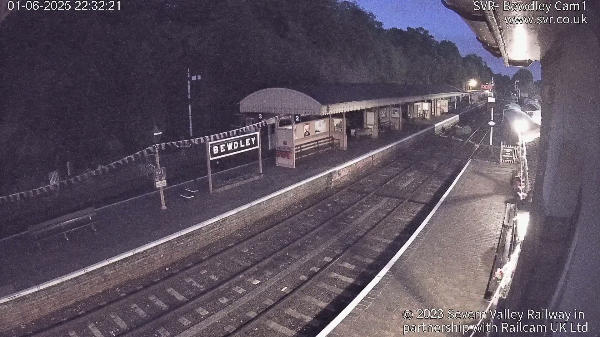 A nighttime view of a train station platform with train tracks and a partially visible train under a dark sky.