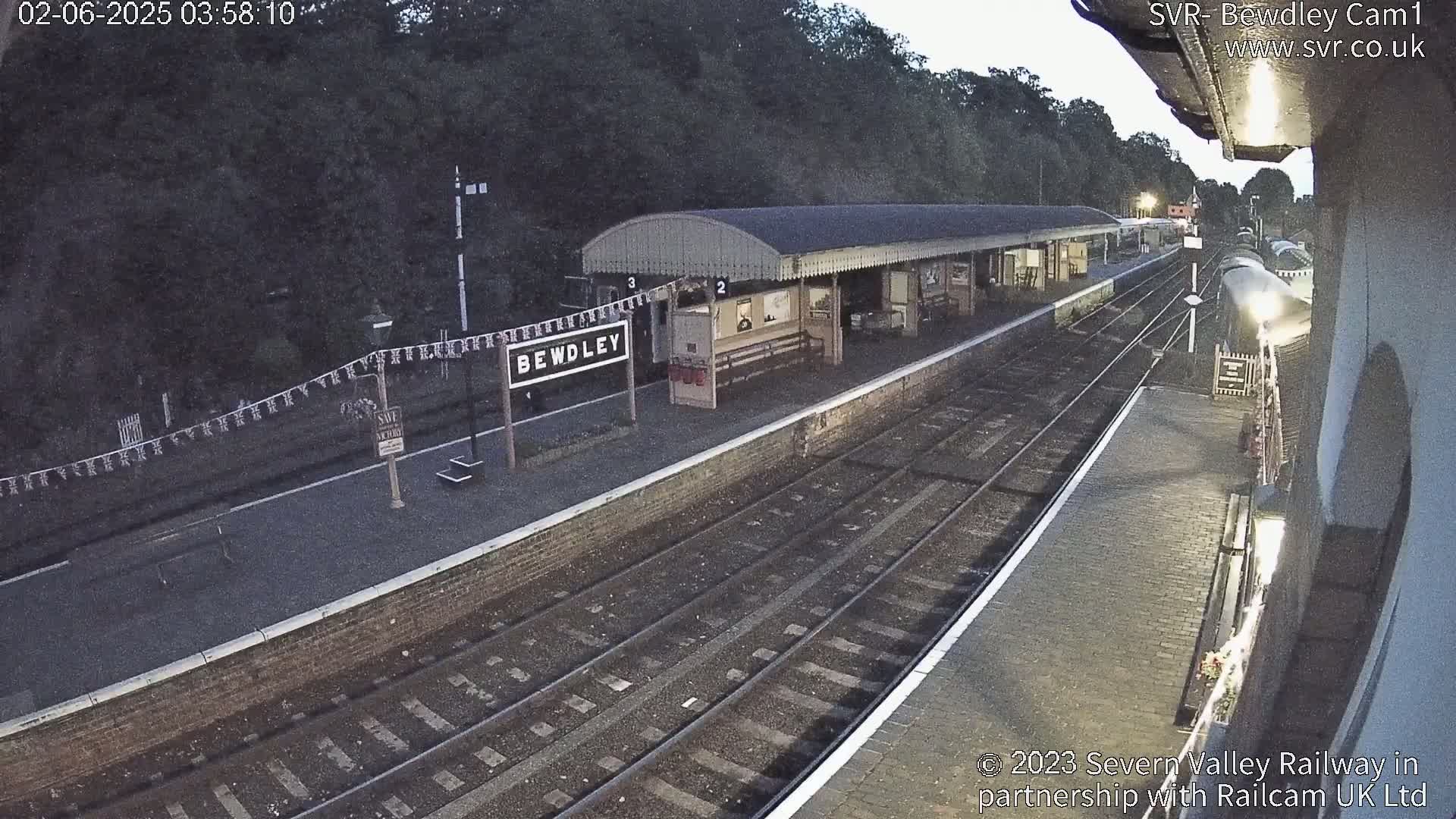 A train sits at a dimly lit railway station platform under a dark, possibly early morning sky.