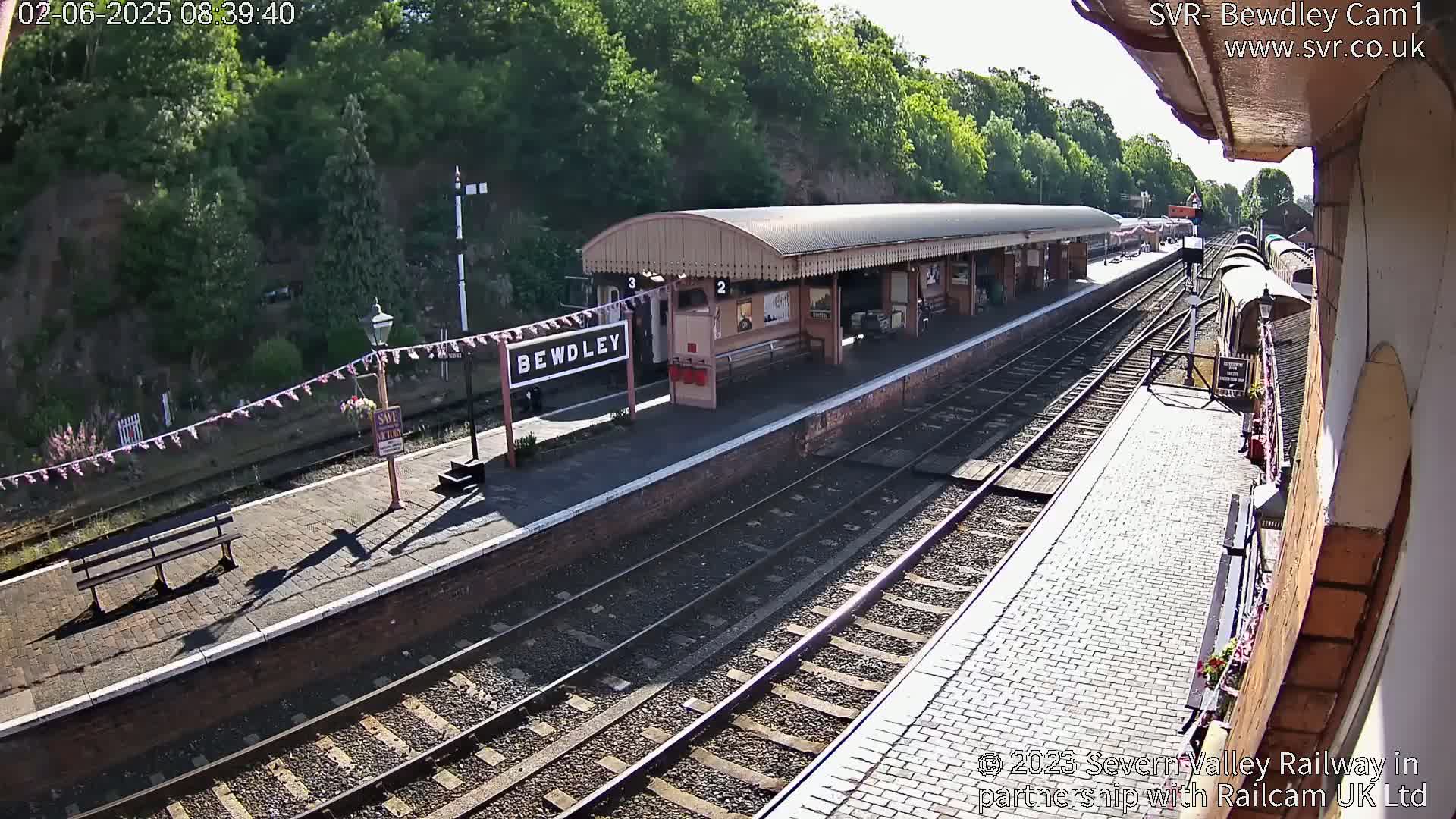 A sunny view of a train station platform with multiple train tracks, a covered waiting area, and a train partially visible in the distance.