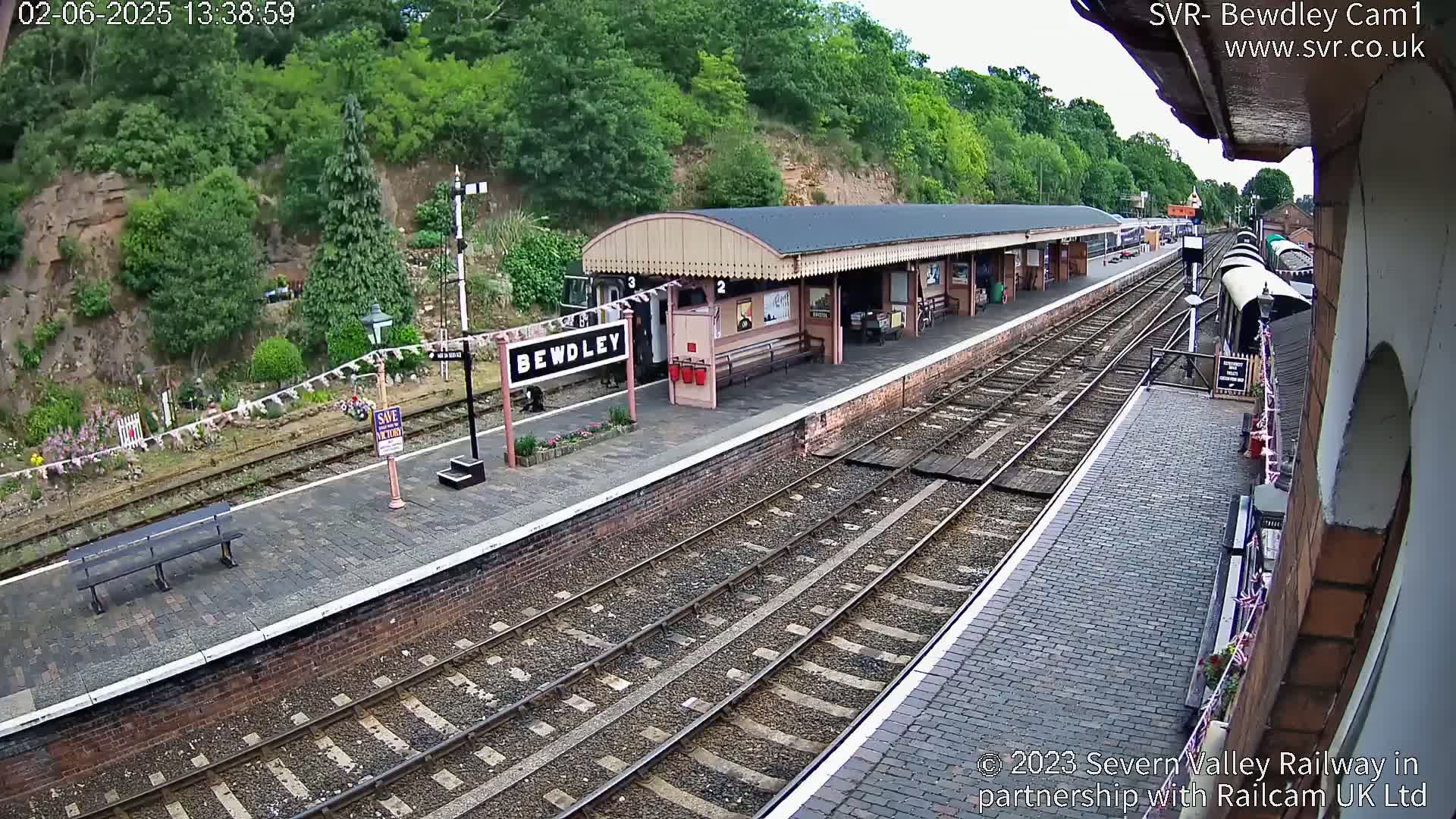 A train station platform with multiple train tracks, a covered waiting area, and lush green hills in the background on a sunny day.