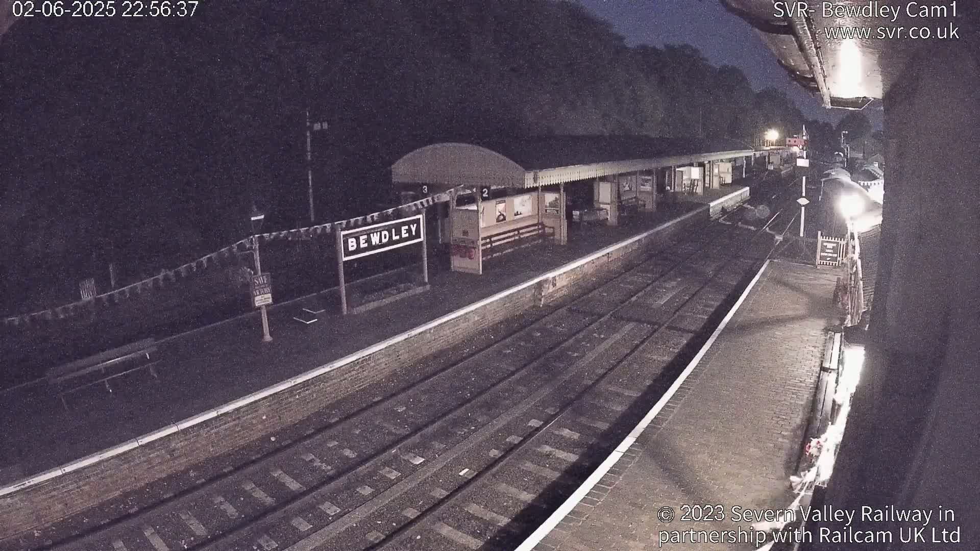 A nighttime view of a train station platform with train tracks, a building, and a partially visible train under dark, nighttime conditions.