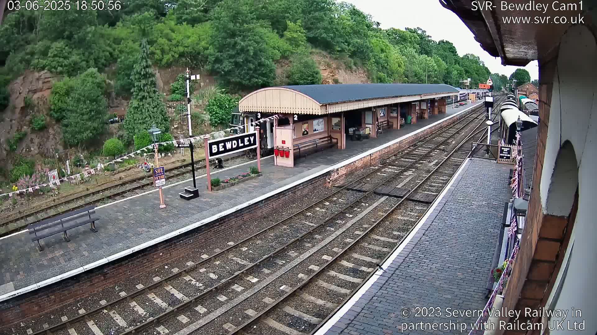 A train station platform with a covered waiting area, multiple train tracks, and lush green hills in the background on an overcast day.