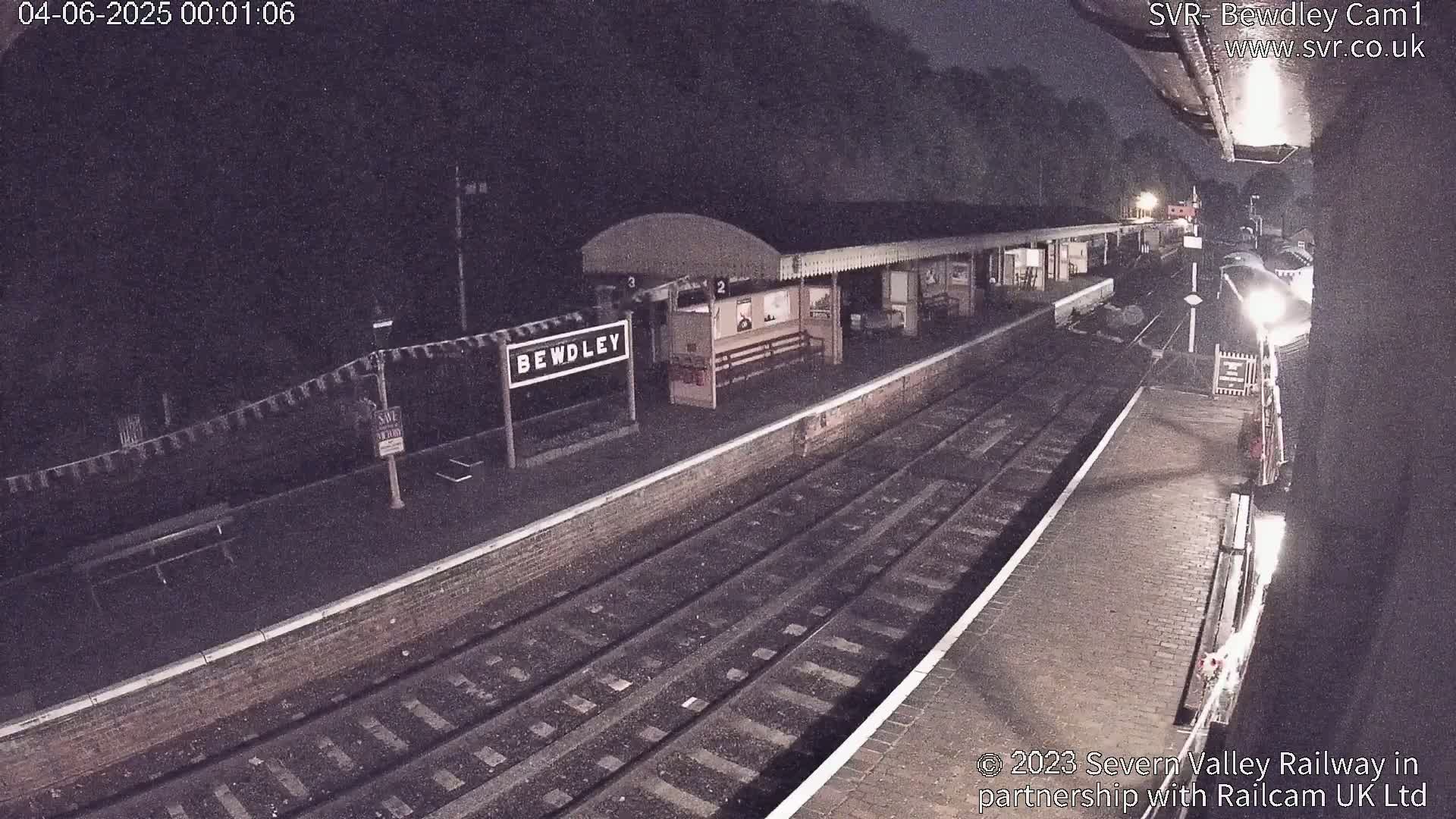 A nighttime view of a train station platform with train tracks, a small station building, and a sign indicating the station's name.