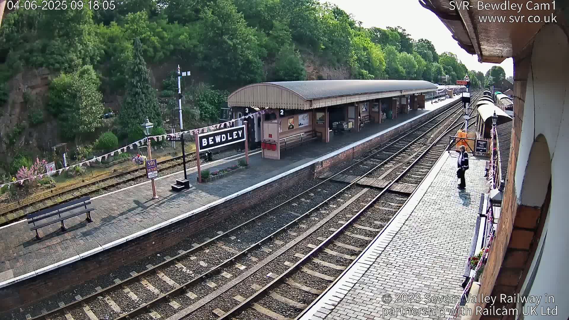 A train station platform with multiple train tracks, a covered waiting area, and a person standing on the platform under a partly sunny sky.