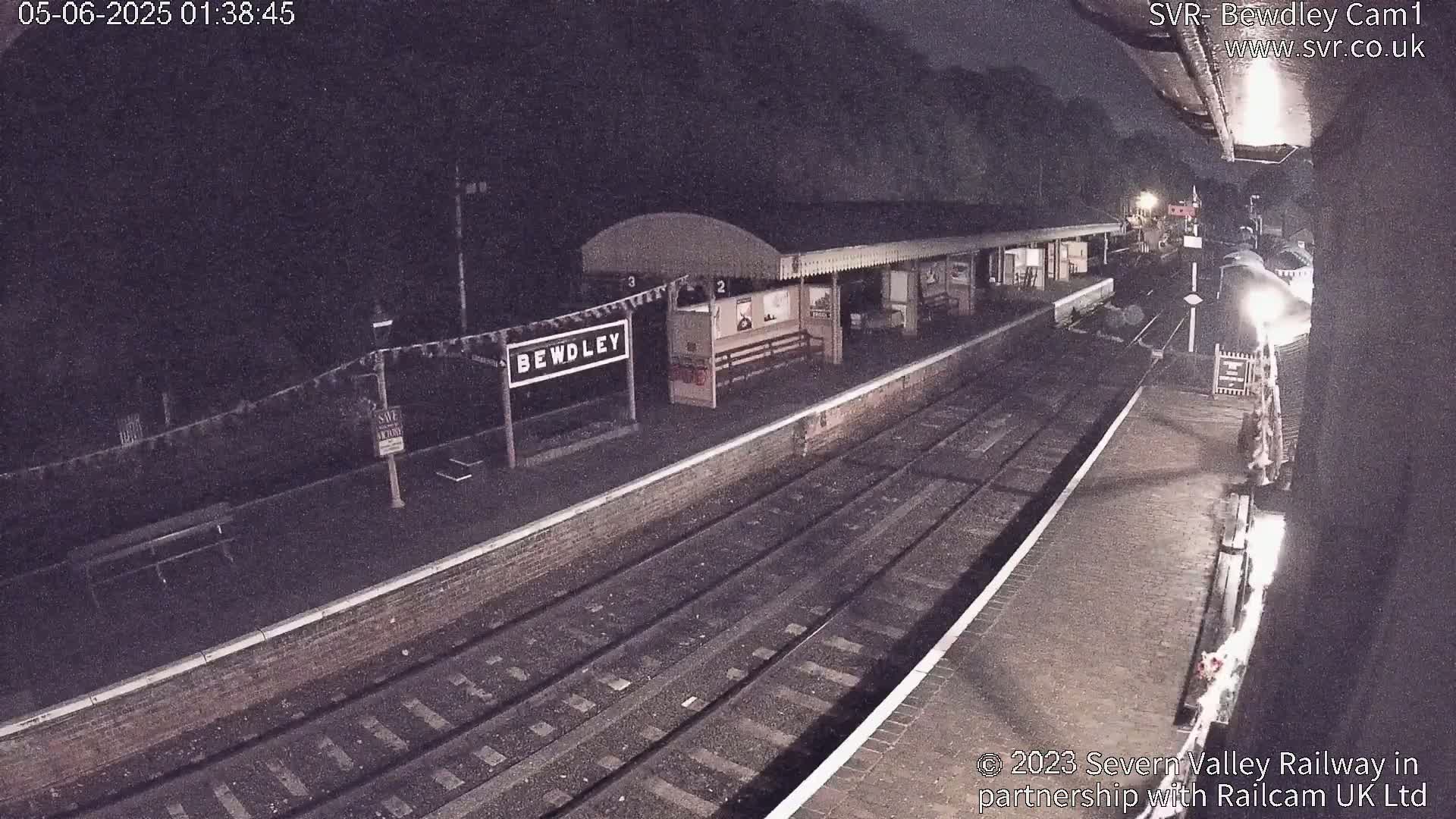 A nighttime view of a train station platform with a train arriving under dark skies.