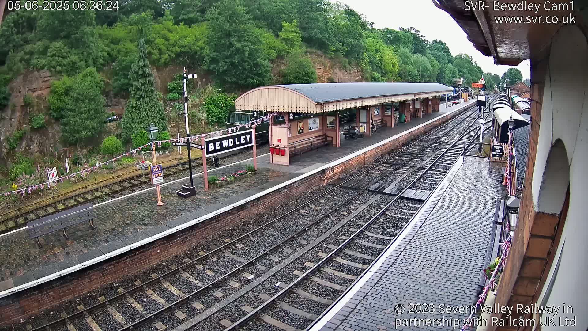 A wet railway station platform with a train partially visible, situated beside a hillside covered in lush greenery.