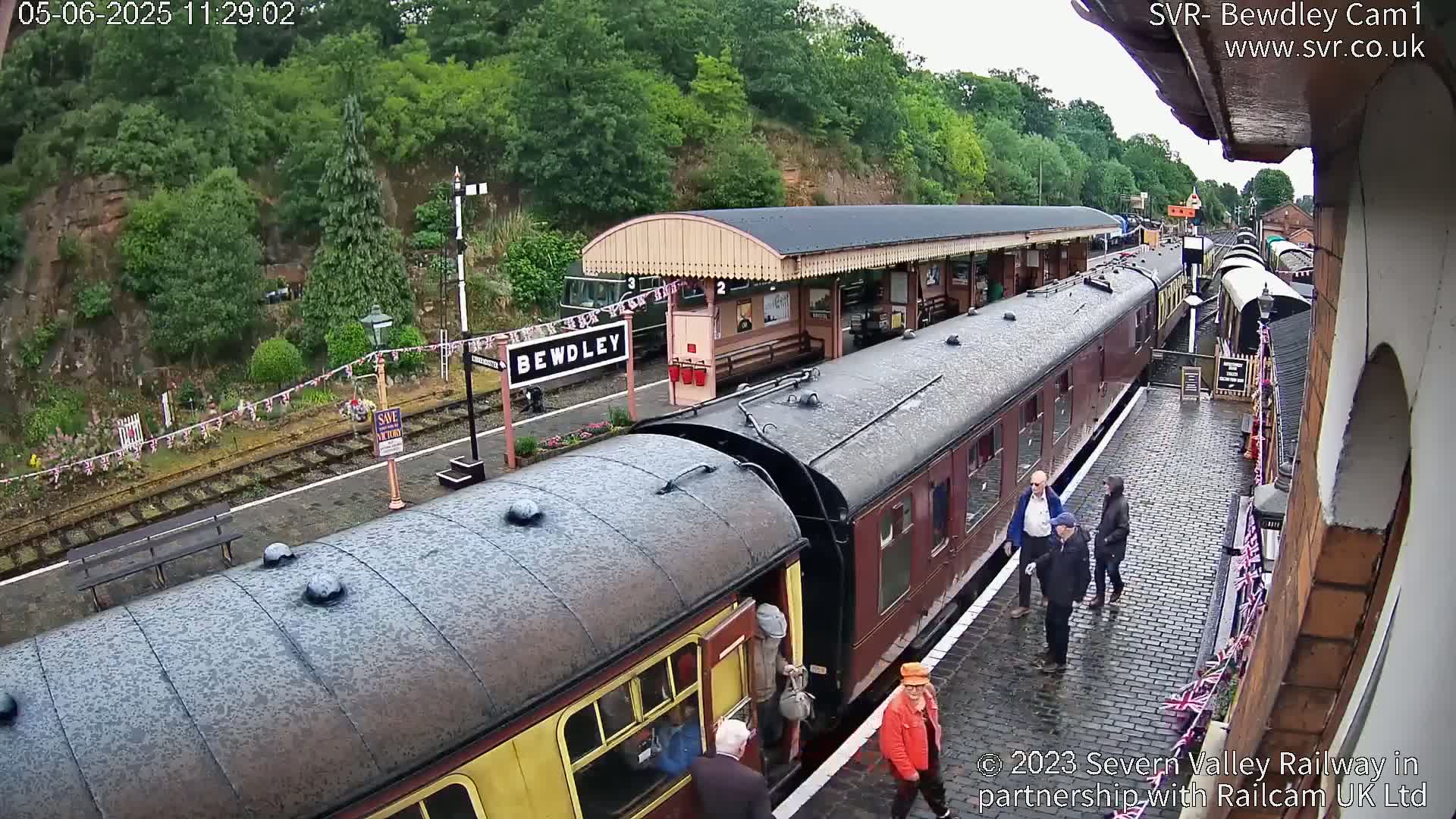 A wet, overcast day at a train station shows several vintage train cars alongside a platform with people waiting and walking nearby.