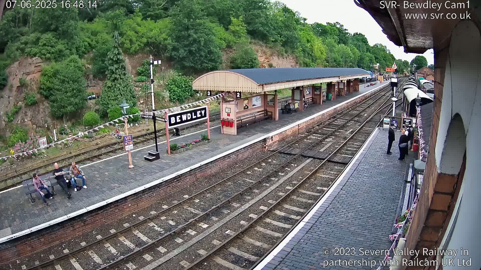 A high-angle view of a train station platform with several people sitting and standing, under a mostly sunny sky.
