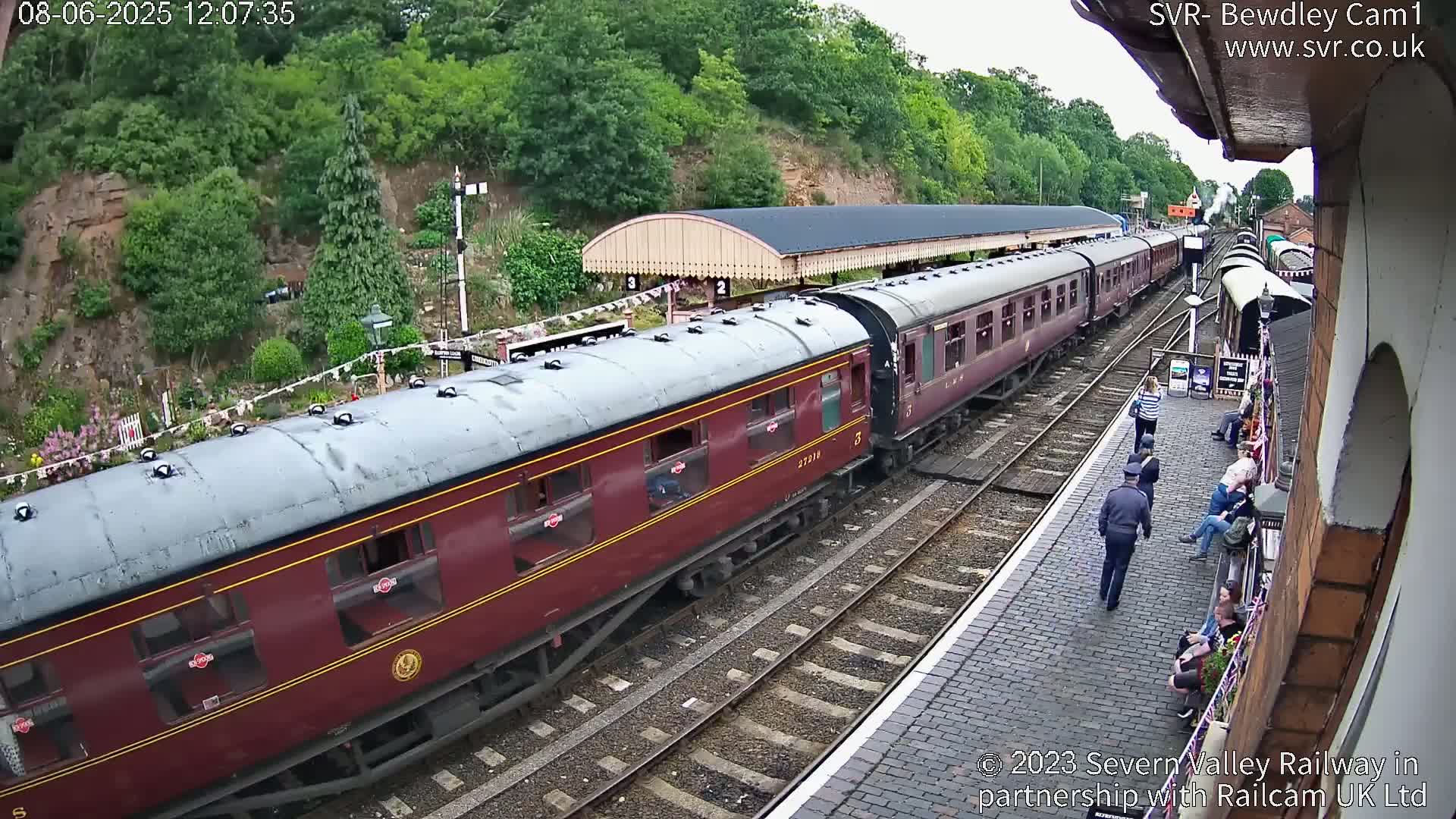 A maroon passenger train is departing a station on a sunny day, with people standing on the platform.