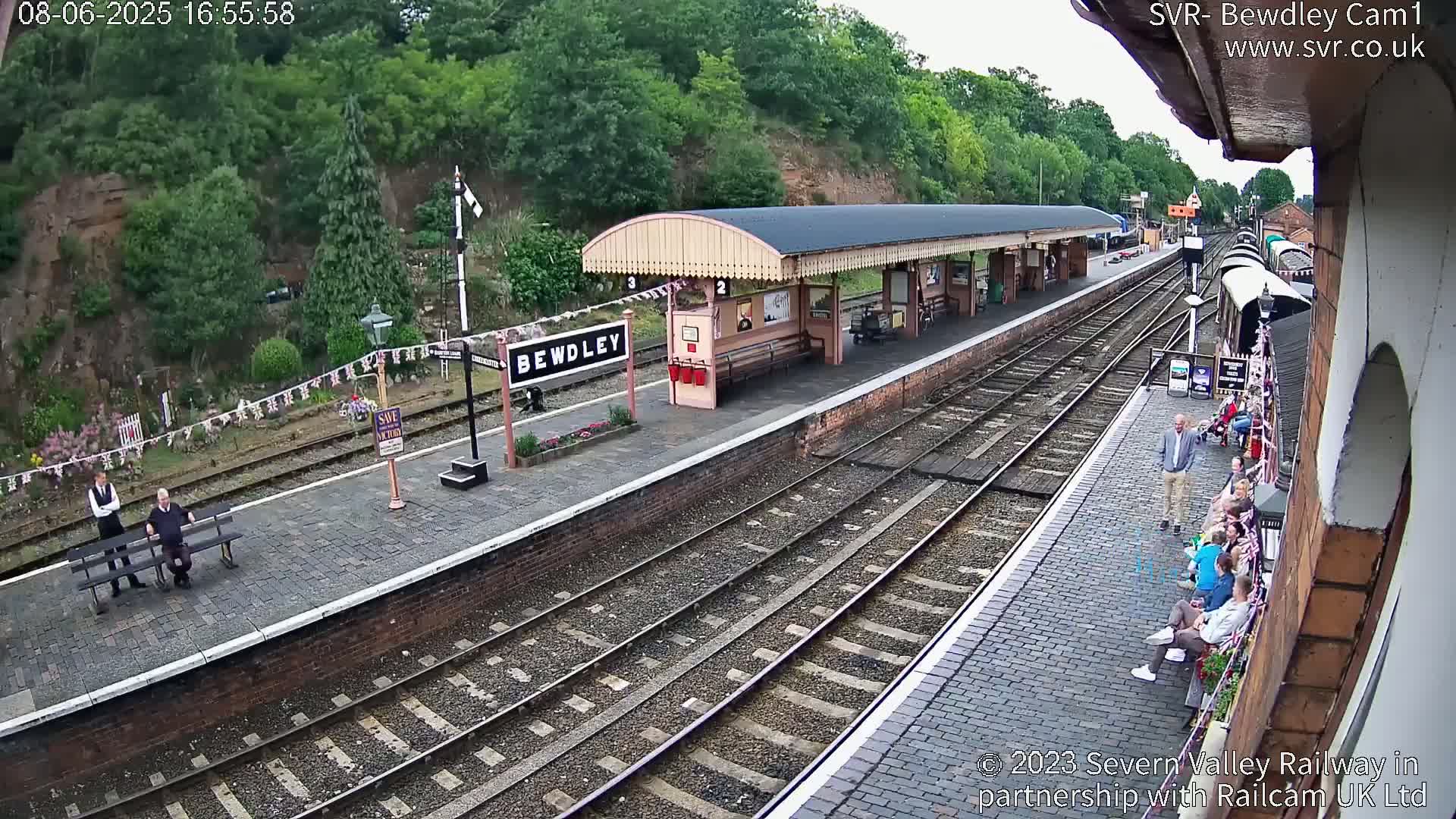 An overcast day at a train station with multiple sets of tracks, a covered platform, and several people waiting.