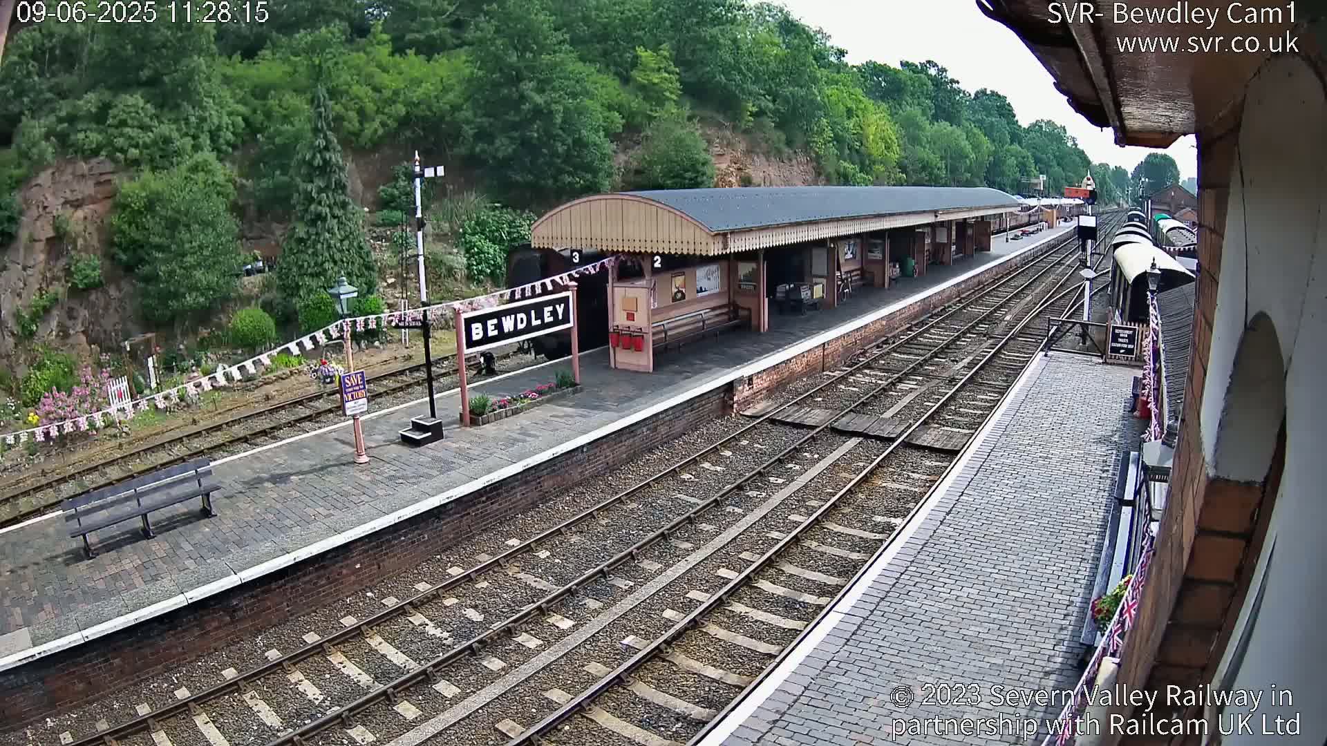 A train station platform with multiple train tracks and a covered waiting area, situated on a hillside on a partly sunny day.