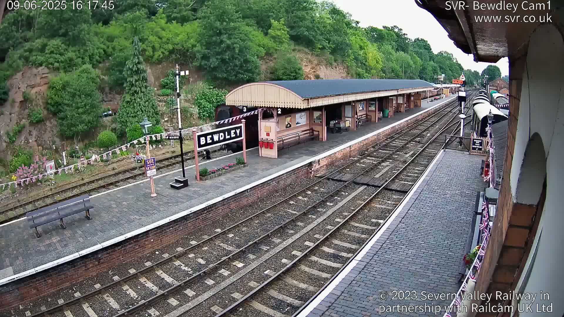 A train station with a covered platform, railway tracks, and a partially visible train, all situated on a hillside under a partly cloudy sky.