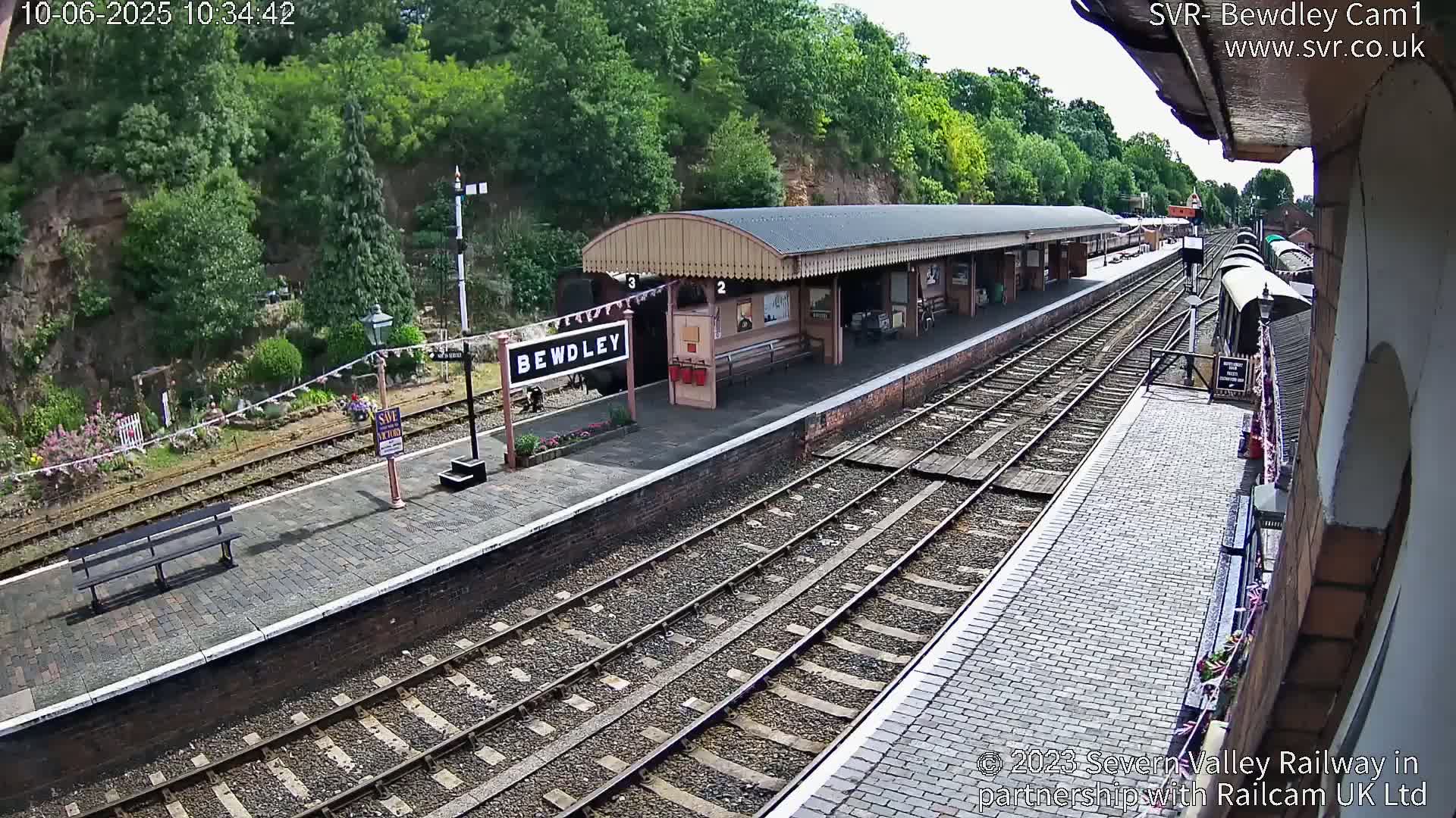 A train station platform with multiple train tracks and a partially visible train, under a sunny sky.