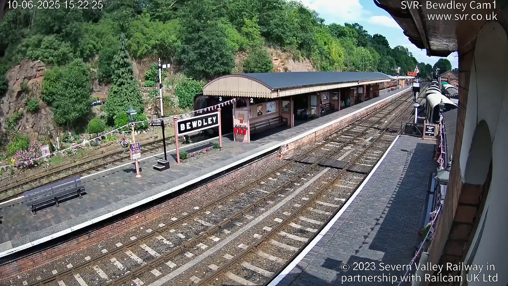 A train station with a covered platform, multiple train tracks, and a train partially visible, situated on a hillside with green trees under a sunny sky.