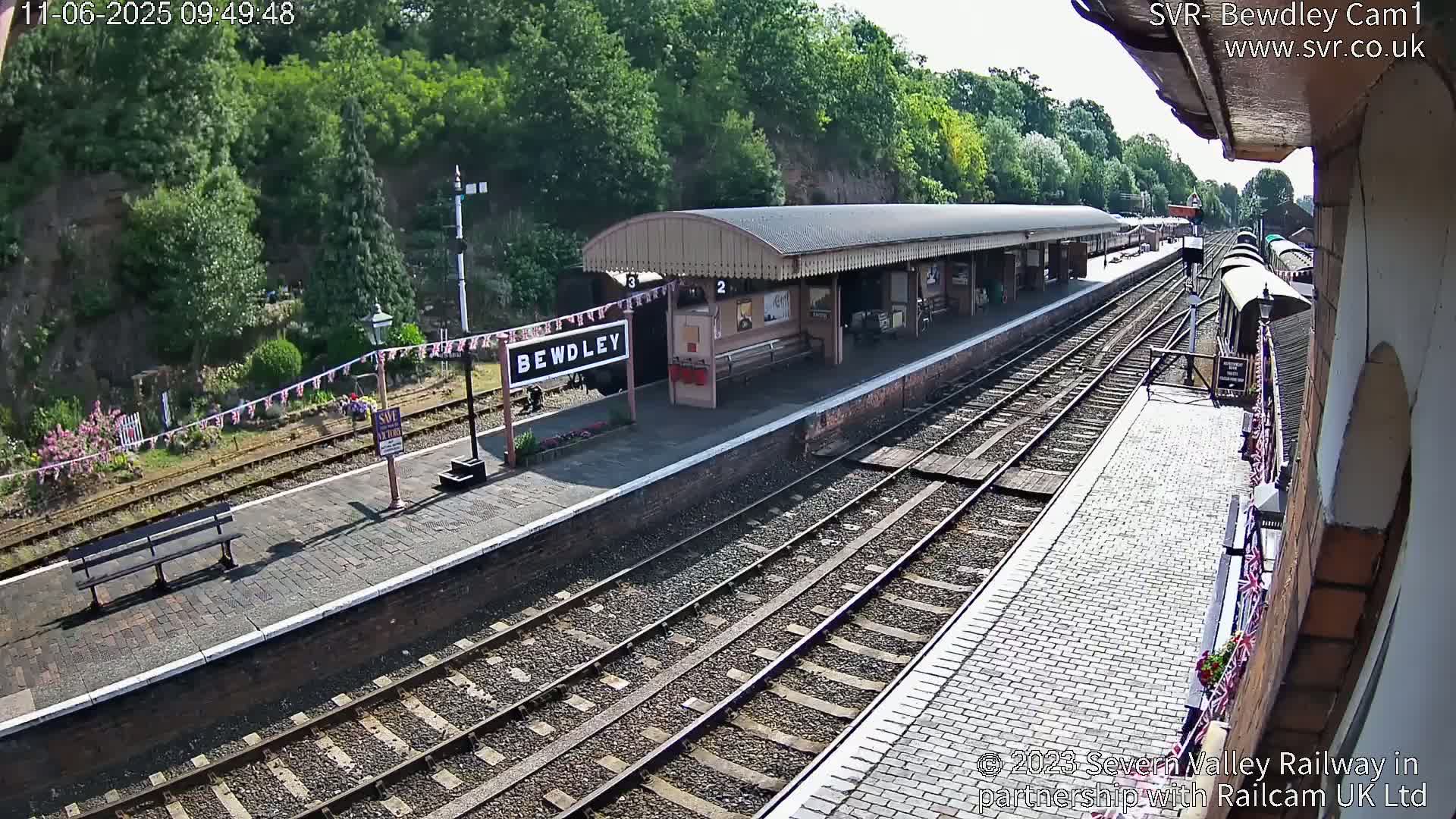 A sunny view of a train station platform with several train tracks, a covered waiting area, and a partially visible train.