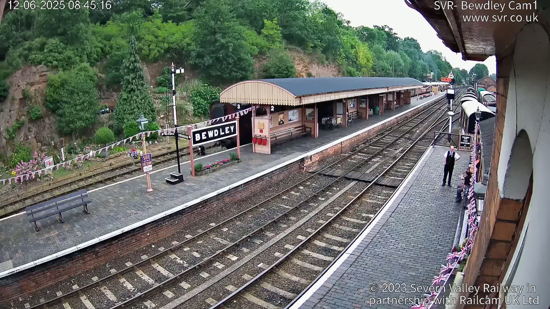 An overhead view of a train station platform with multiple train tracks, a partially visible train, and a person standing on the platform under overcast skies.