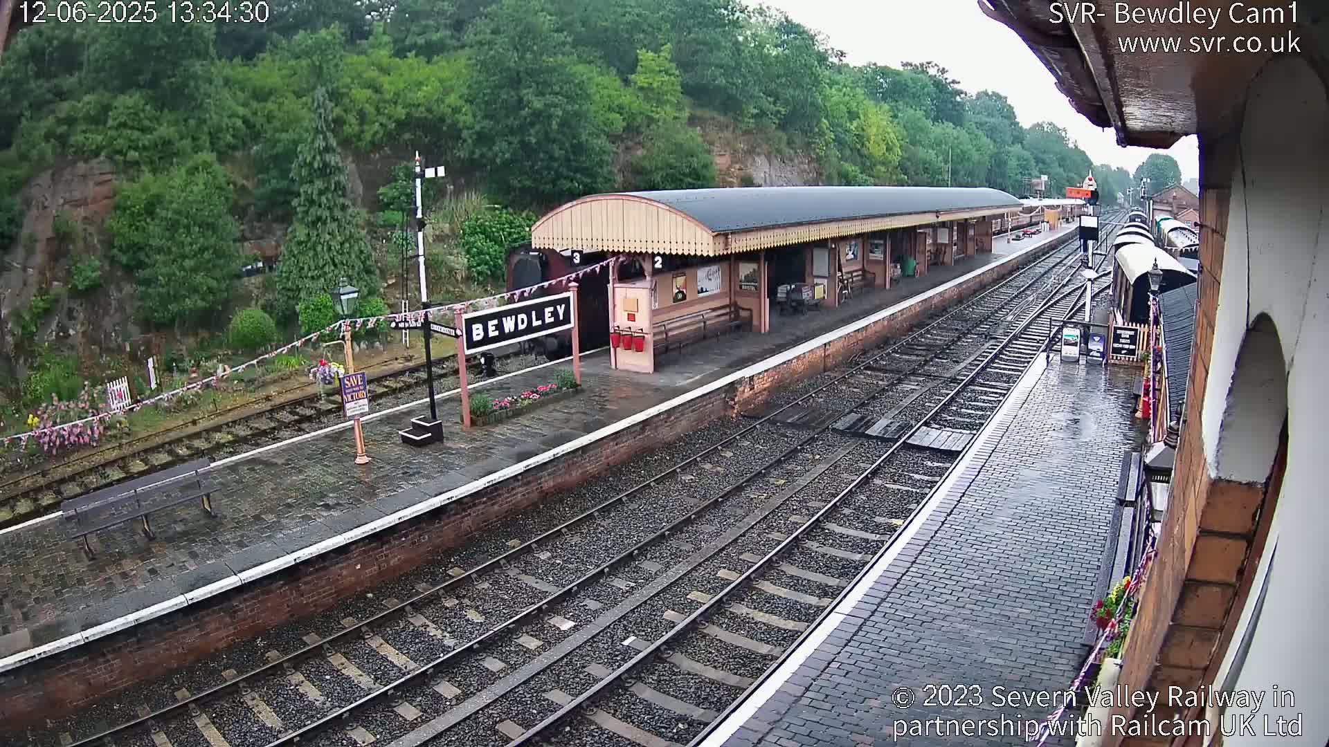 A rain-slicked railway station platform with multiple train tracks, a covered waiting area, and a train partially visible.