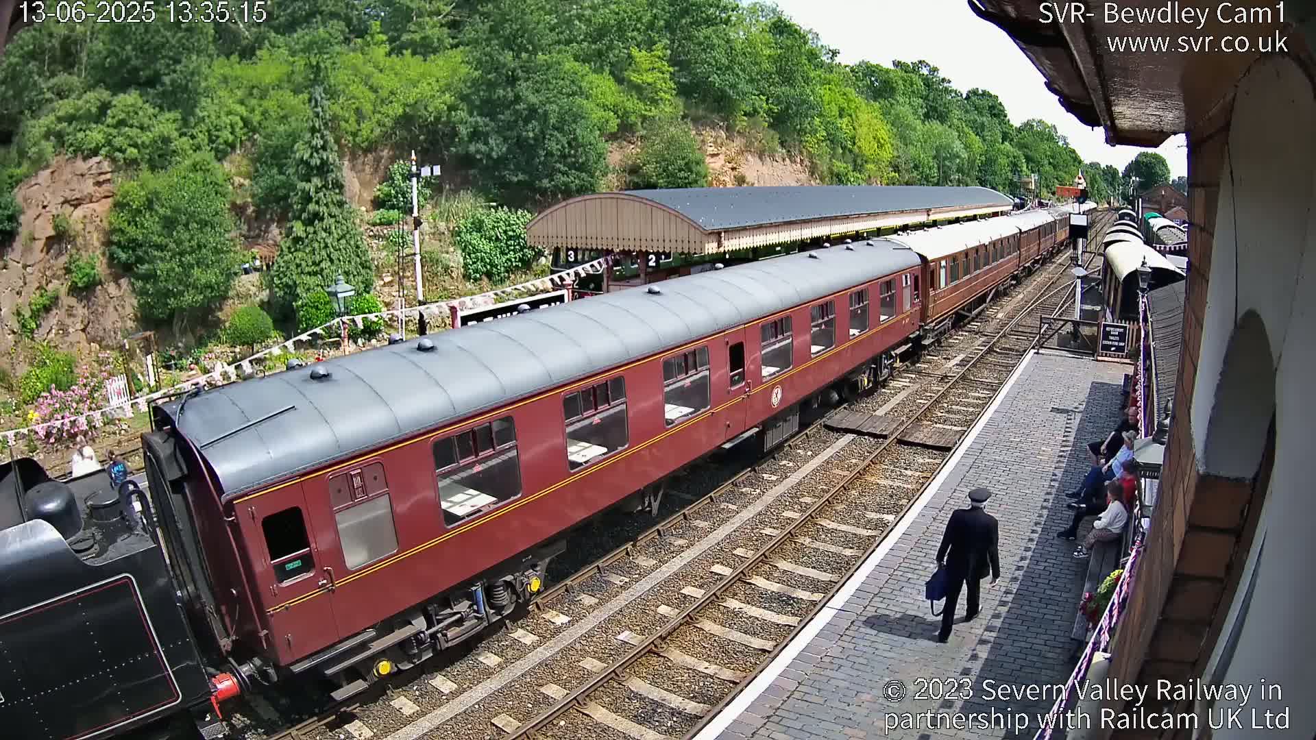 A maroon passenger train sits at a railway station on a sunny day, with a few people waiting on the platform and one walking away.