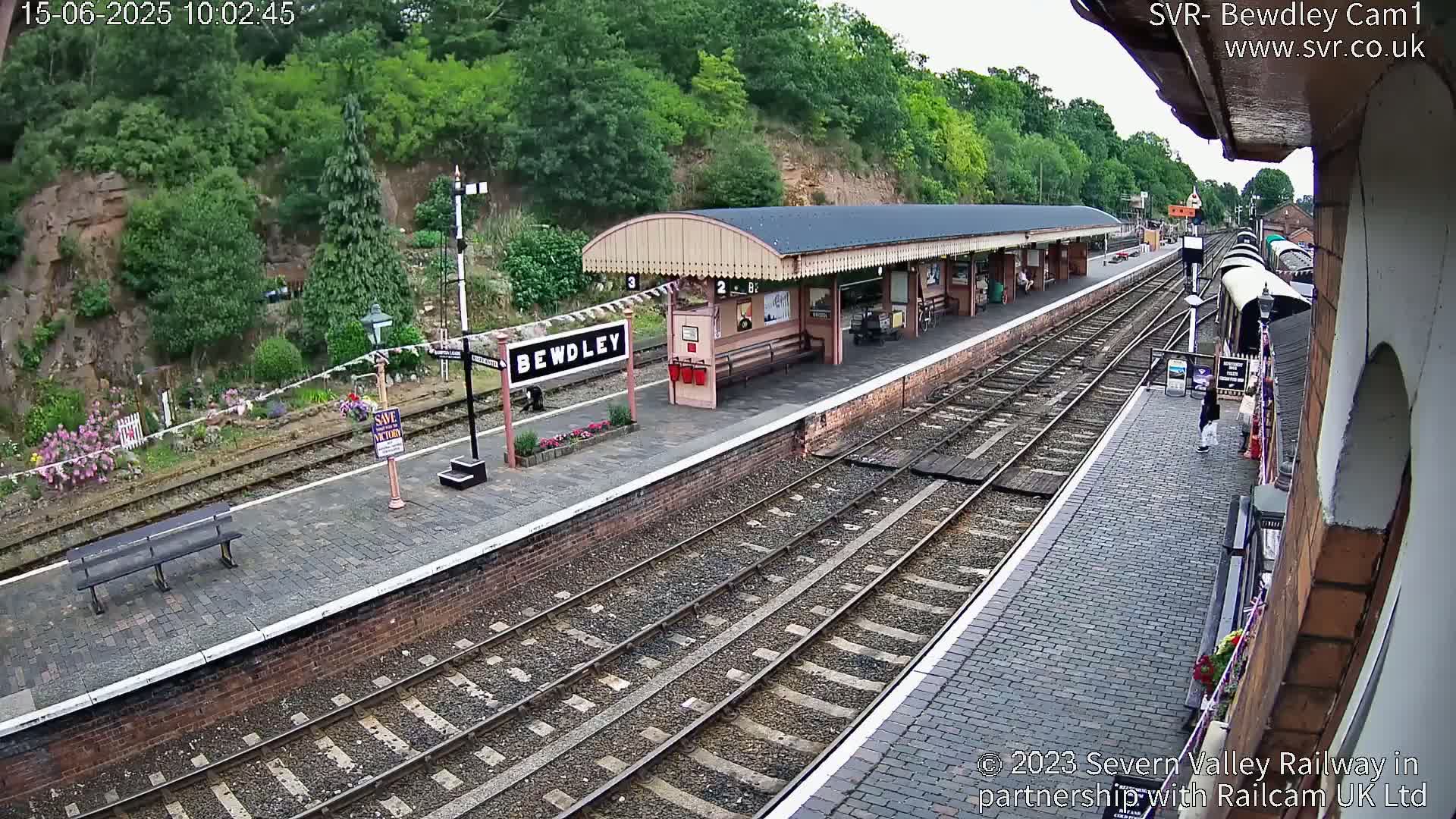 A train station platform with multiple train tracks, a covered waiting area, and lush greenery on a partly cloudy day.