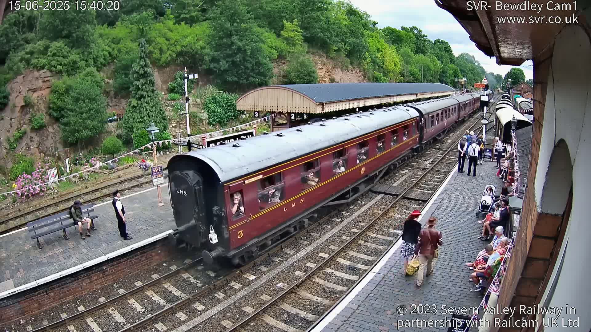A maroon passenger train is arriving at a railway station on a partly sunny day, with people waiting on the platform and a few sitting on benches.