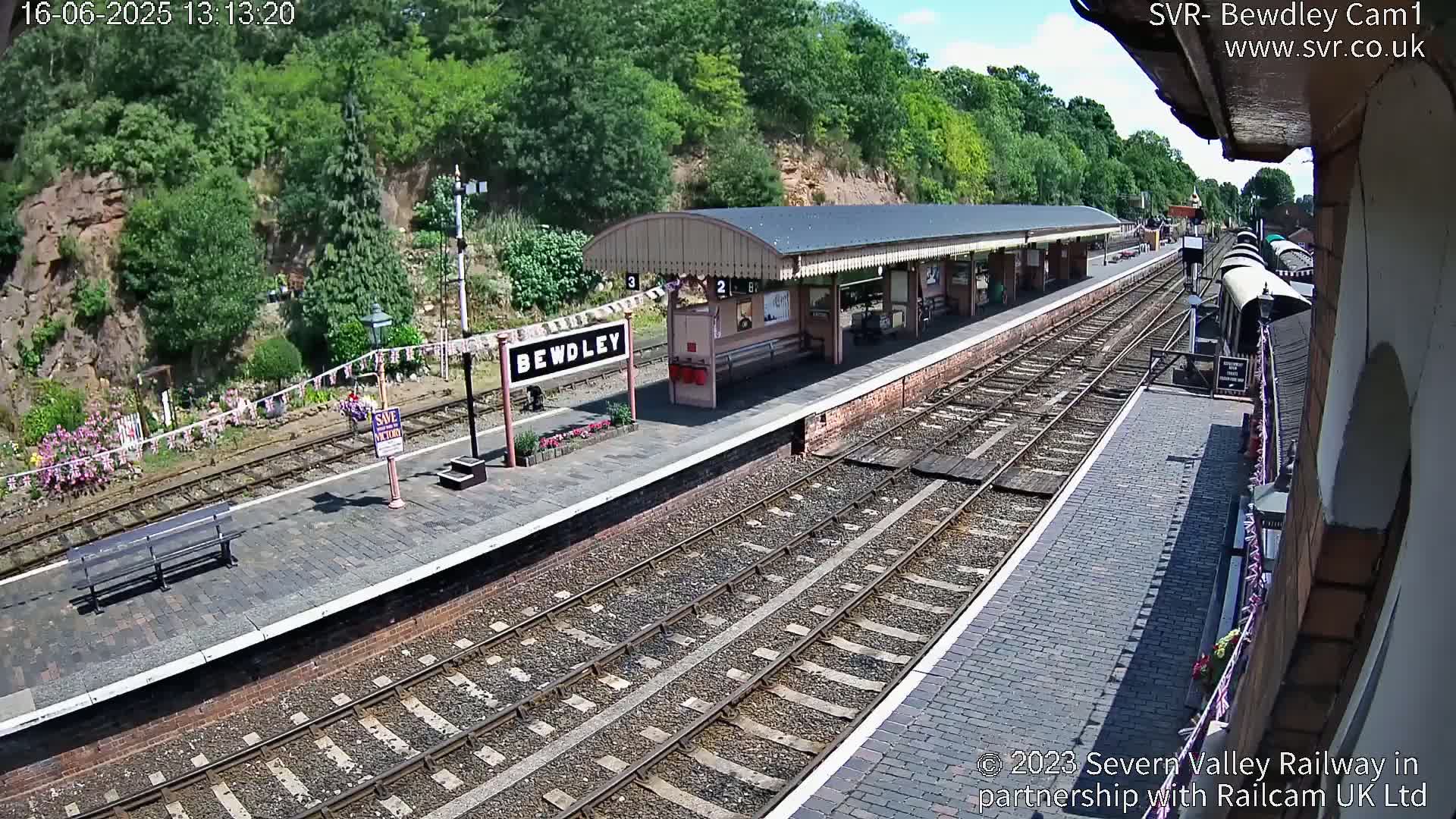 A railway station platform with a covered waiting area, multiple train tracks, and a background of lush green hills under a sunny sky.