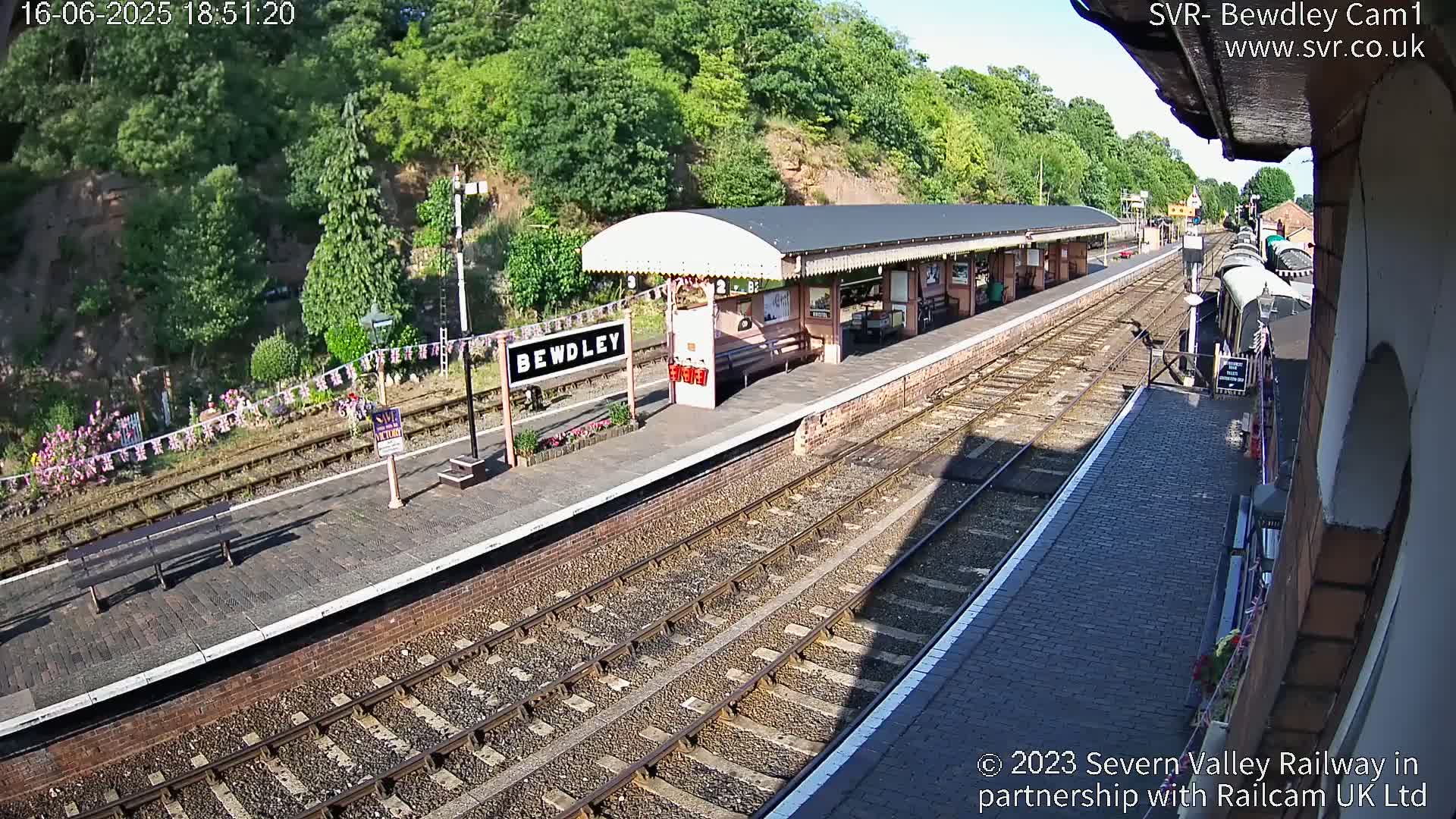 A sunny day at a train station with a covered platform, train tracks, and a train partially visible in the distance.