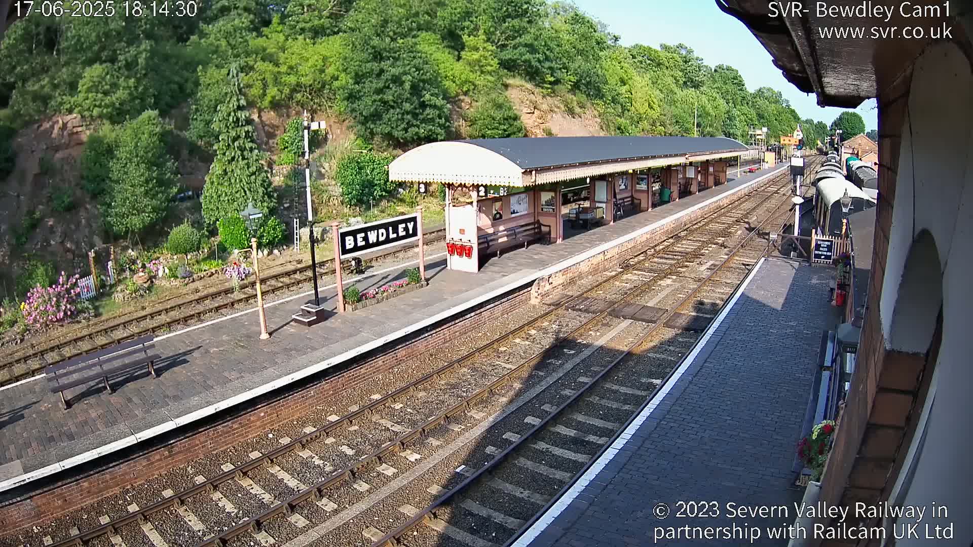 A sunny view of a train station platform with railway tracks, a covered waiting area, and a train car partially visible in the background.